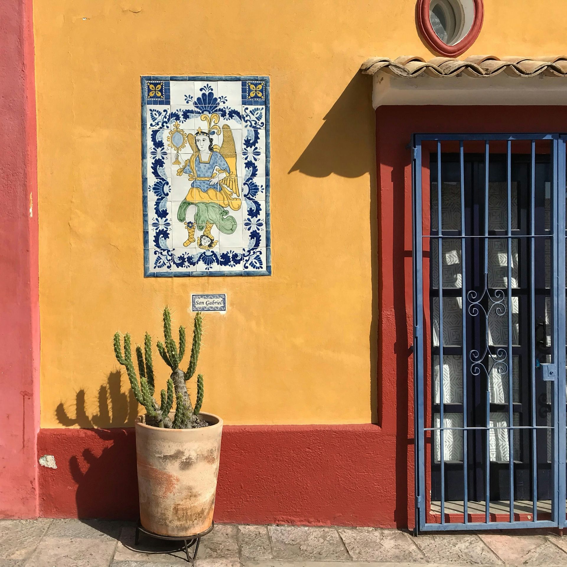 Yellow and red exterior wall with a grilled door, a ceramic tile with a Hindu figure, and a potted cactus in Oaxaca, Mexico.