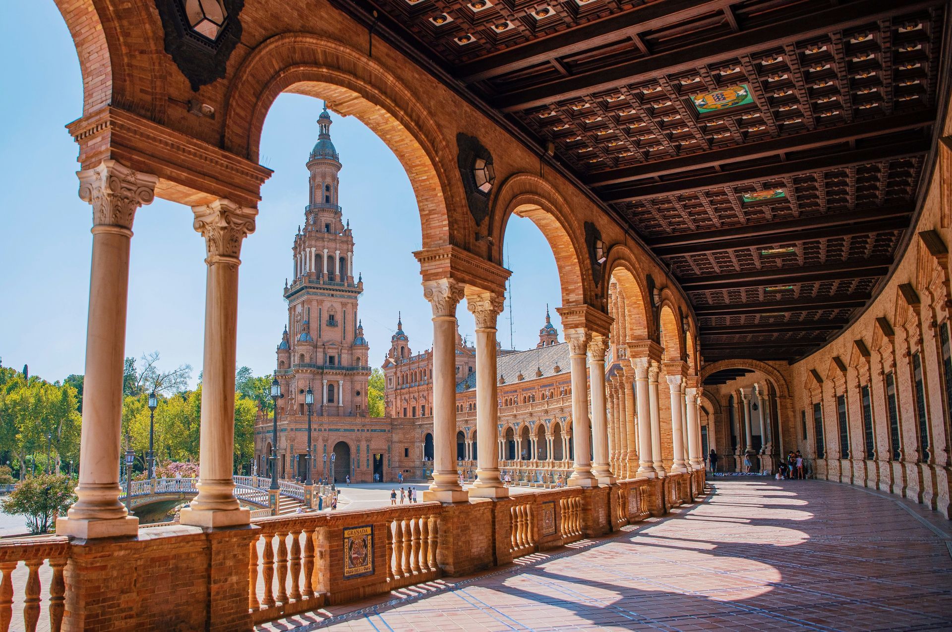 A long arched walkway with view of a tall tower in Seville, Spain, under a clear blue sky.