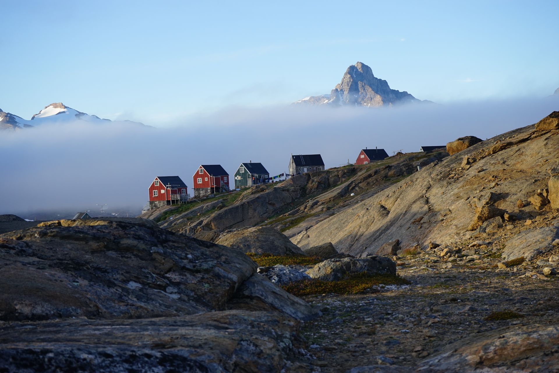 Red and blue houses on a rocky hill, fog rolling below, mountain peak in the distance in Tasiilaq, Greenland.