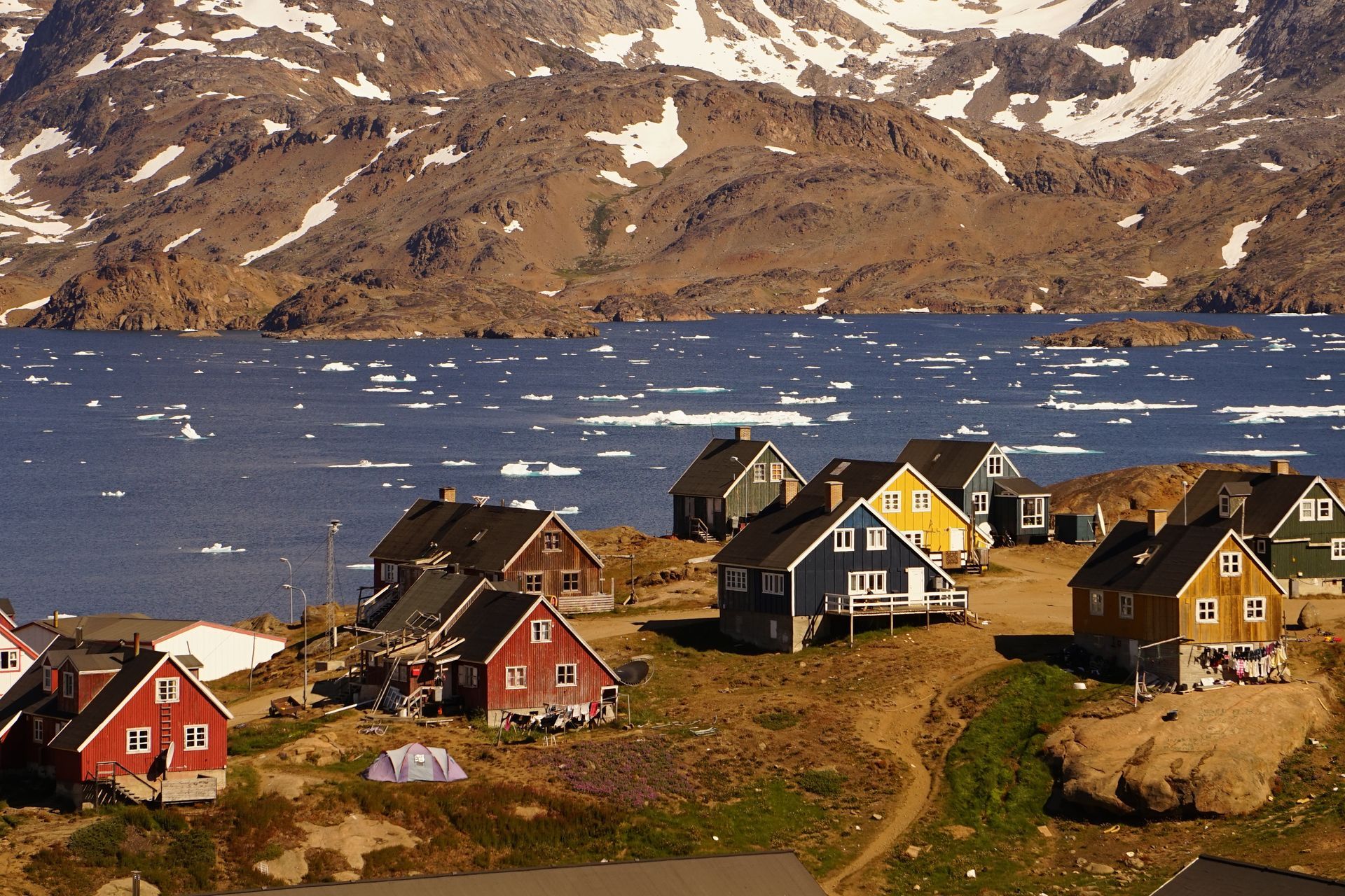 Colorful houses on a hillside overlooking a body of water with ice, mountains in the background in Tasiilaq, Greenland.