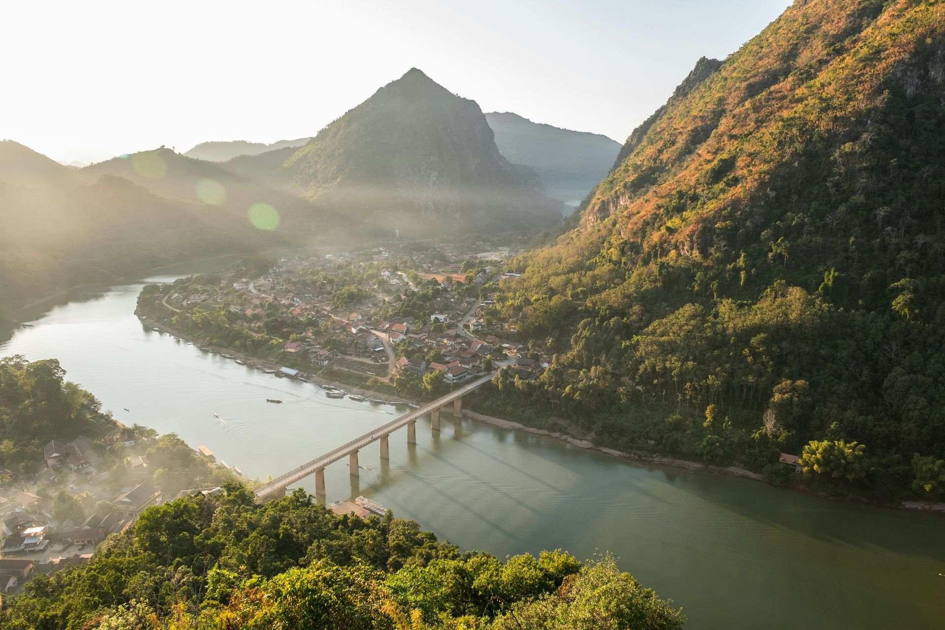 Bridge over a river with the town of Nong Khiaw nestled between mountains; bright sunlight in Laos.