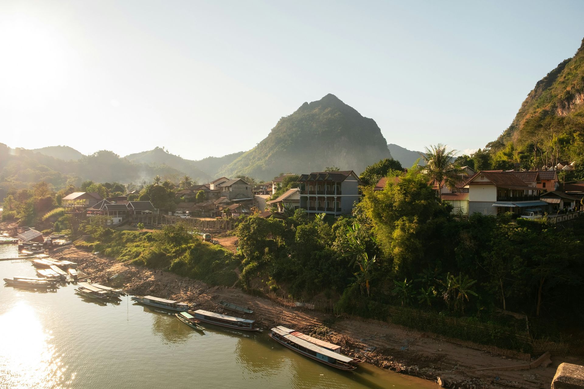 Town of Nong Khiaw nestled by river and mountain, bathed in warm sunlight. Boats line the riverbank in Laos.