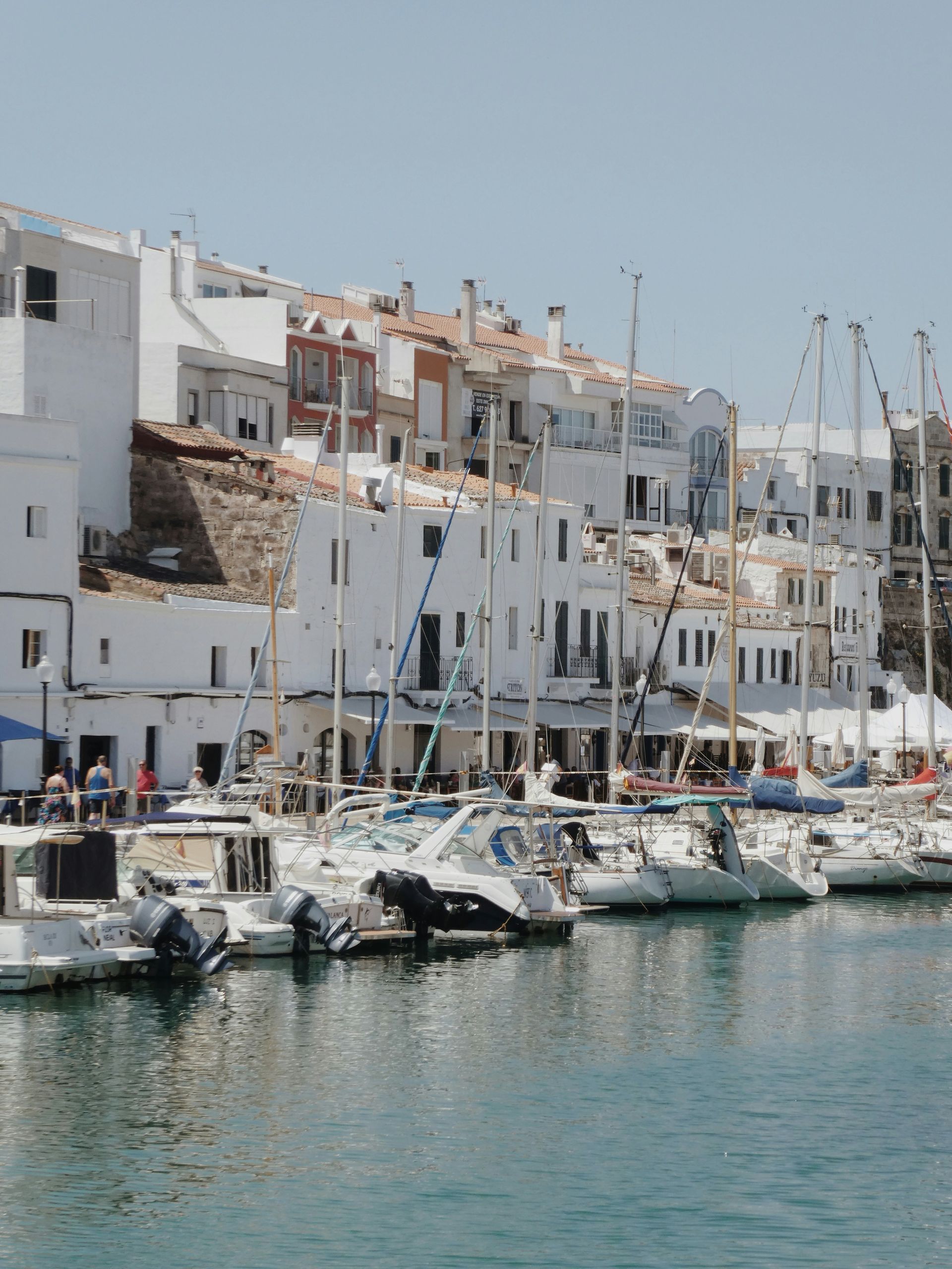 Boats docked at a harbor lined with white buildings, turquoise water in Menorca, Spain.