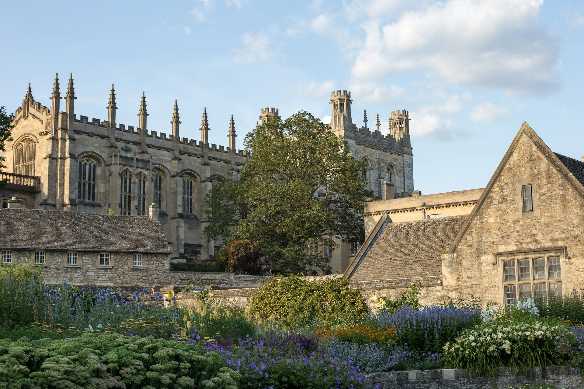 Stone building with tall windows, surrounded by a garden of flowers, under a blue sky at Oxford University, England.