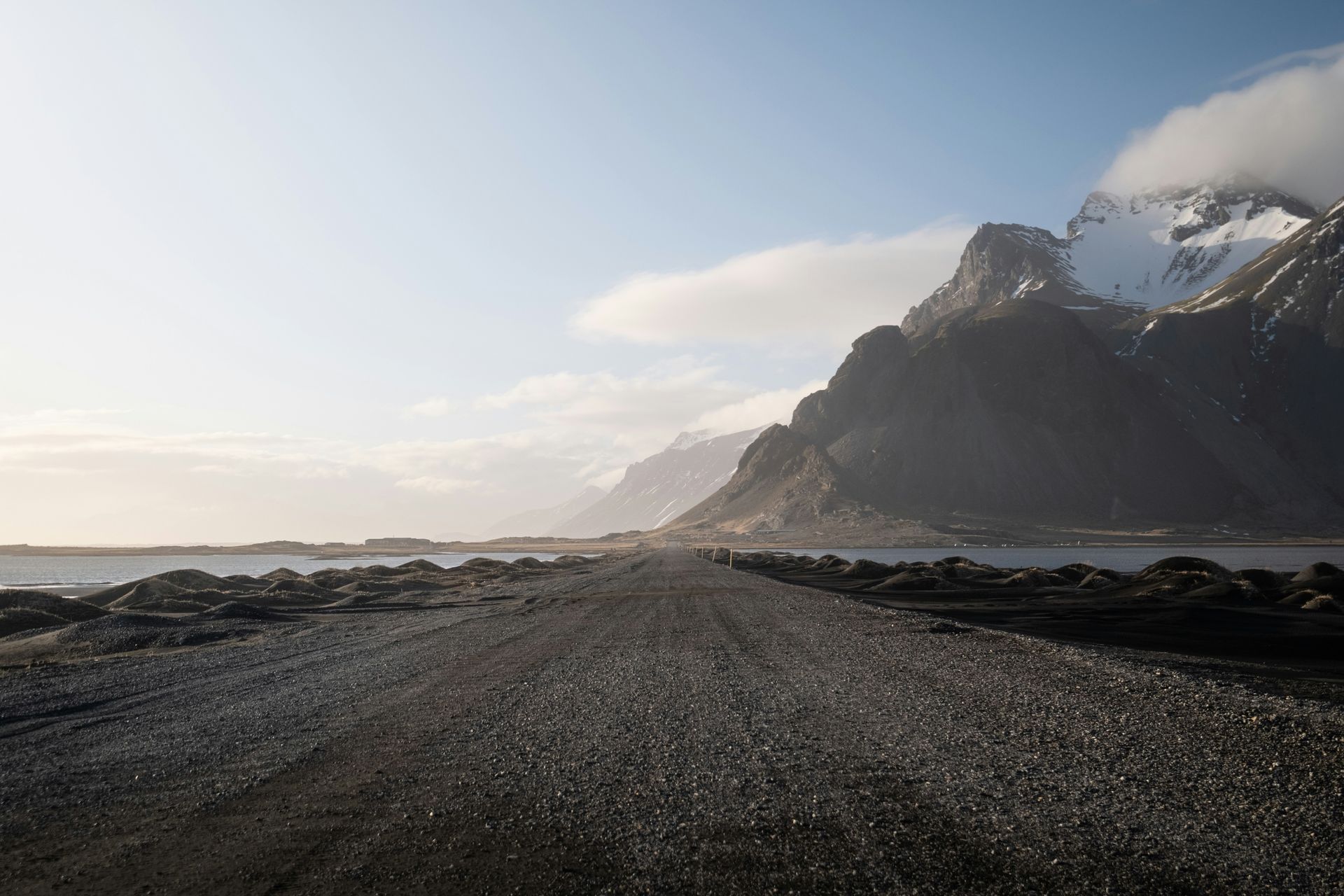 Gravel road leads toward snow-capped mountains under a cloudy sky, near a body of water in Stokksnes, Iceland.