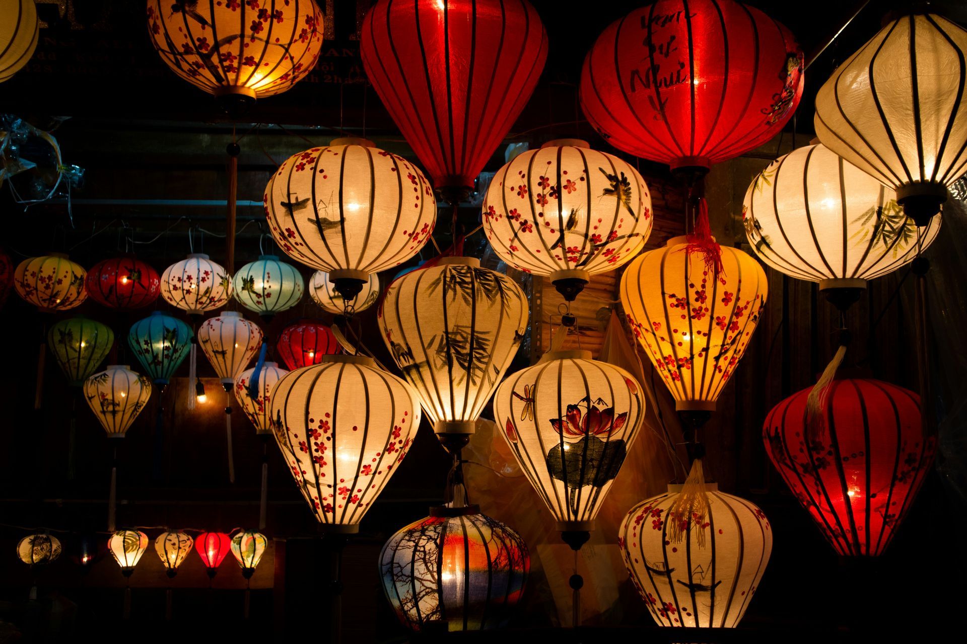 Colorful lanterns, various shapes and sizes, hanging in a dark setting, illuminated with warm light in Hội An, Vietnam.