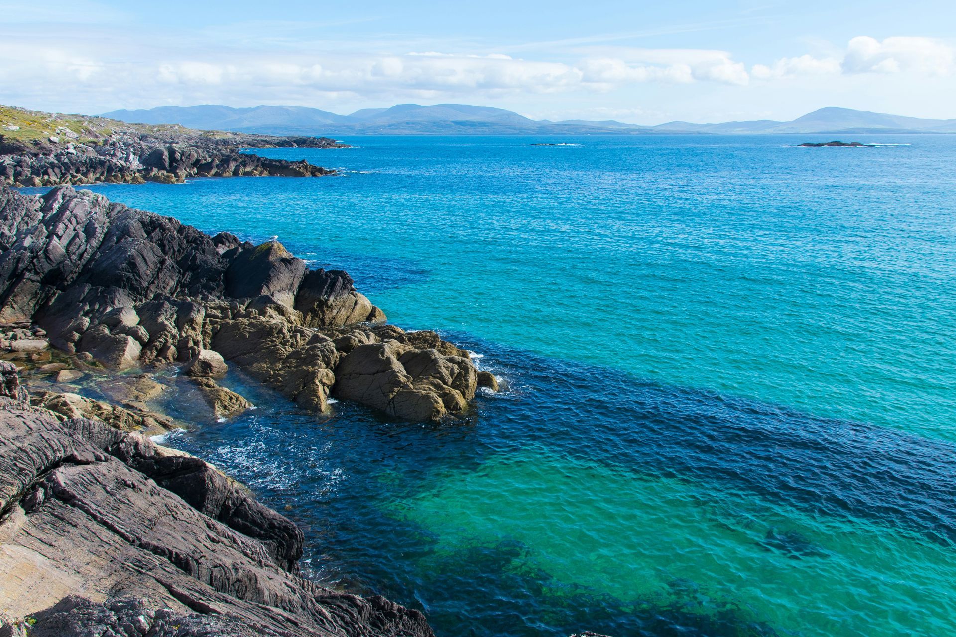 Rocky coastline with turquoise and dark blue water, mountains in the distance on the Ring of Kerry, Ireland.