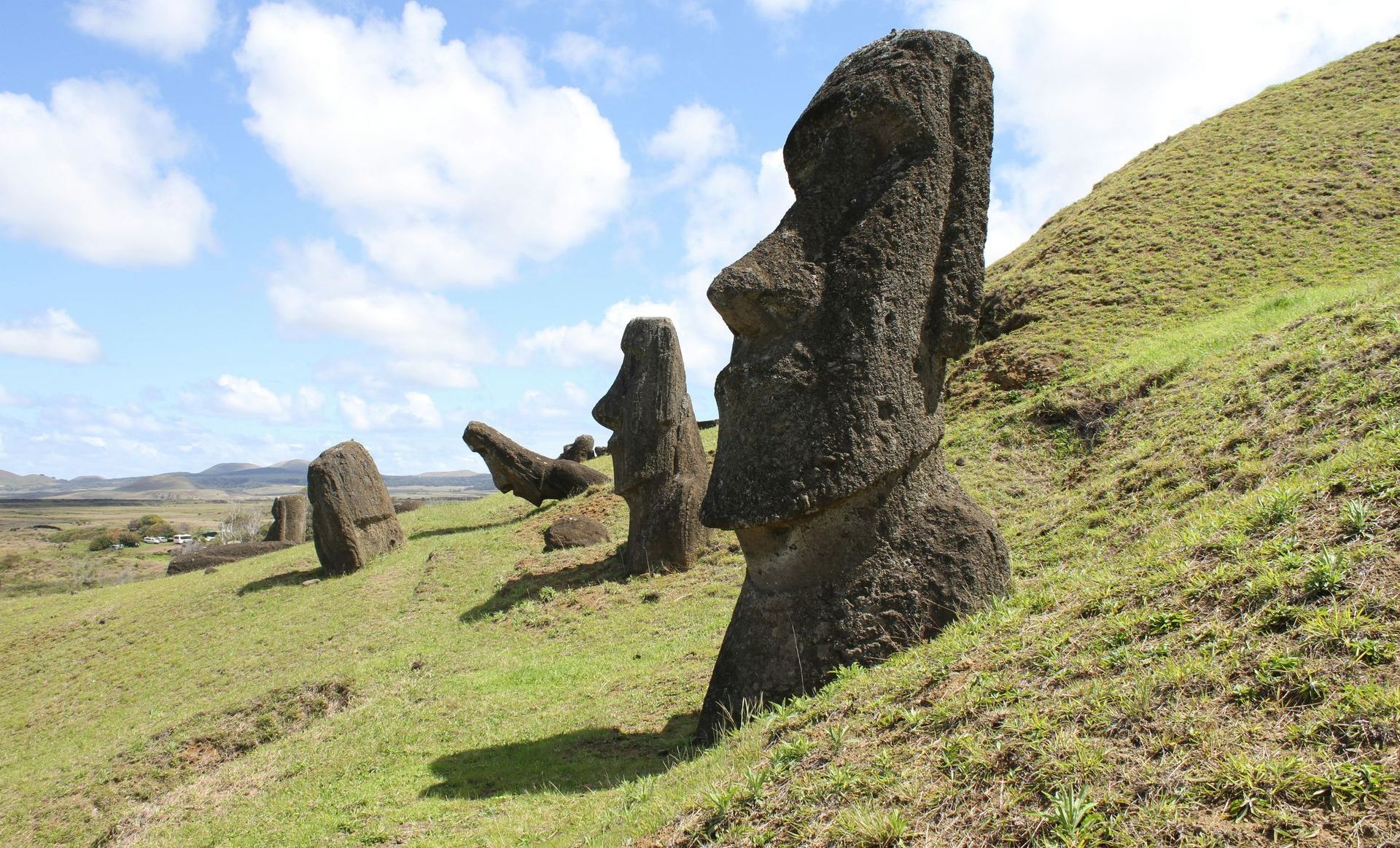 Moai statues on a grassy hill, Easter Island, under a blue sky with clouds.