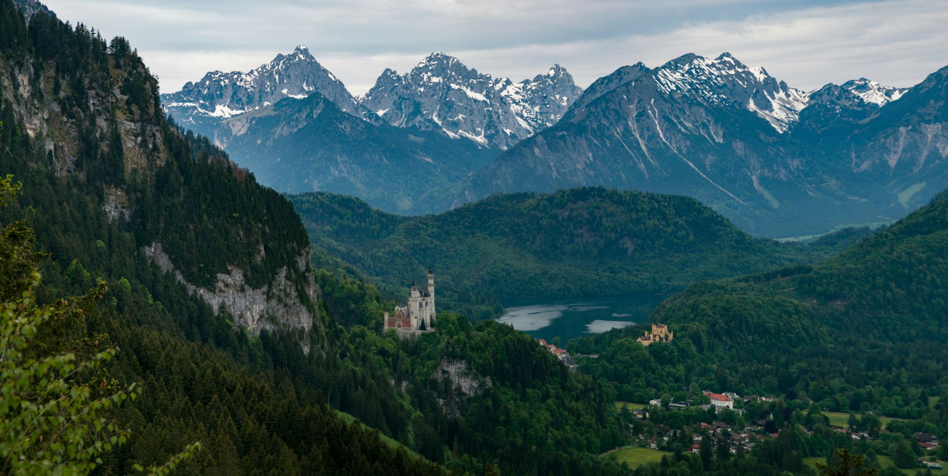 Bavarian Alps with snow-capped peaks tower above a green valley with a castle and village in Germany.