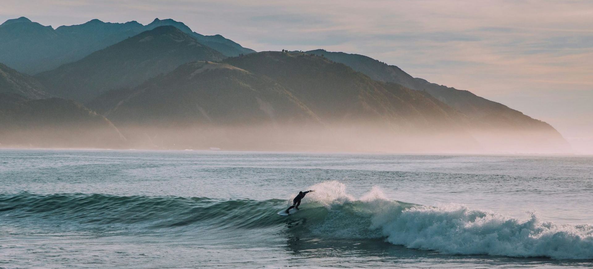A wooden path going through a grassy field with mountains in the background in New Zealand.
