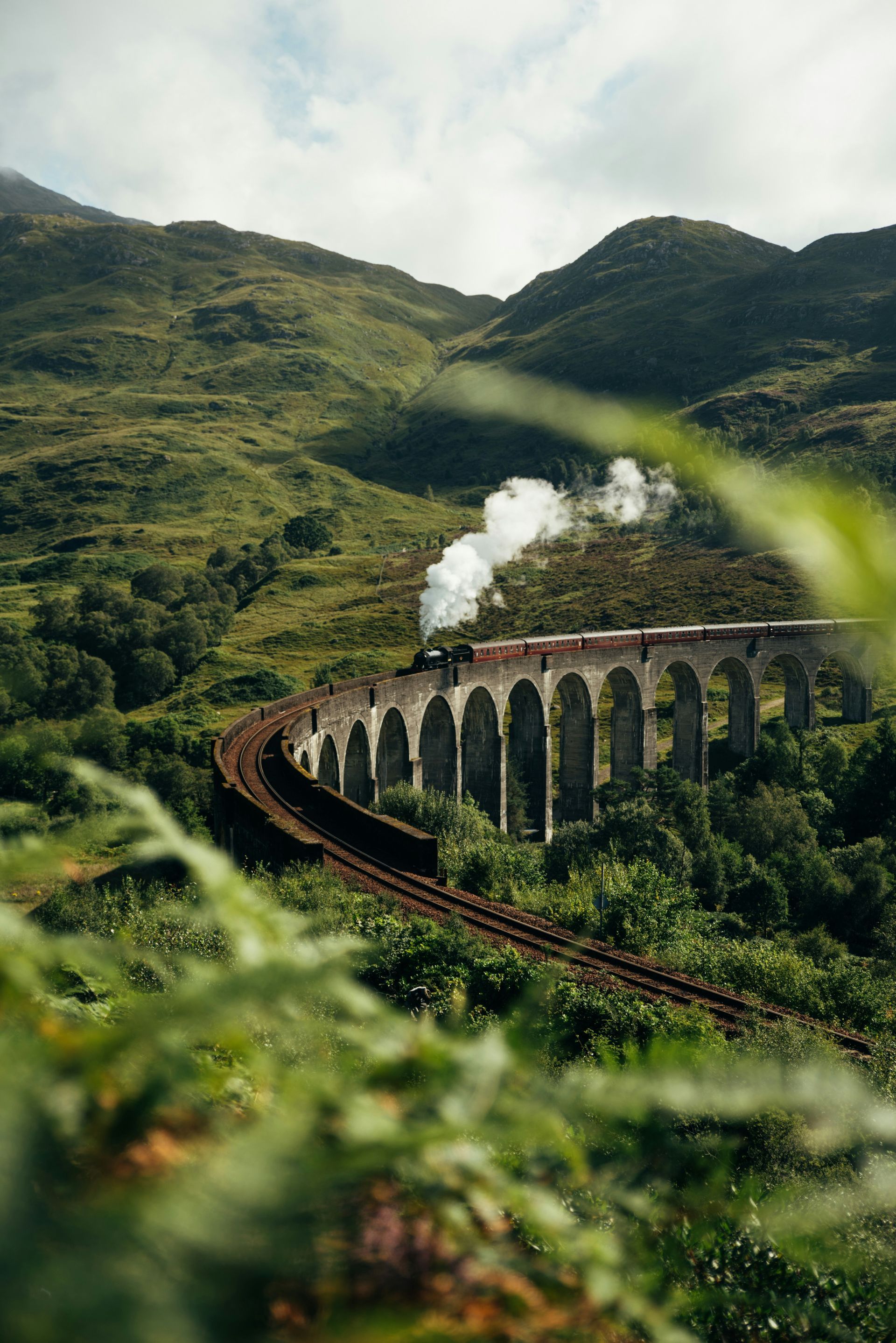 Train on the arched Glenfinnan Viaduct bridge surrounded by green hills in Scotland.