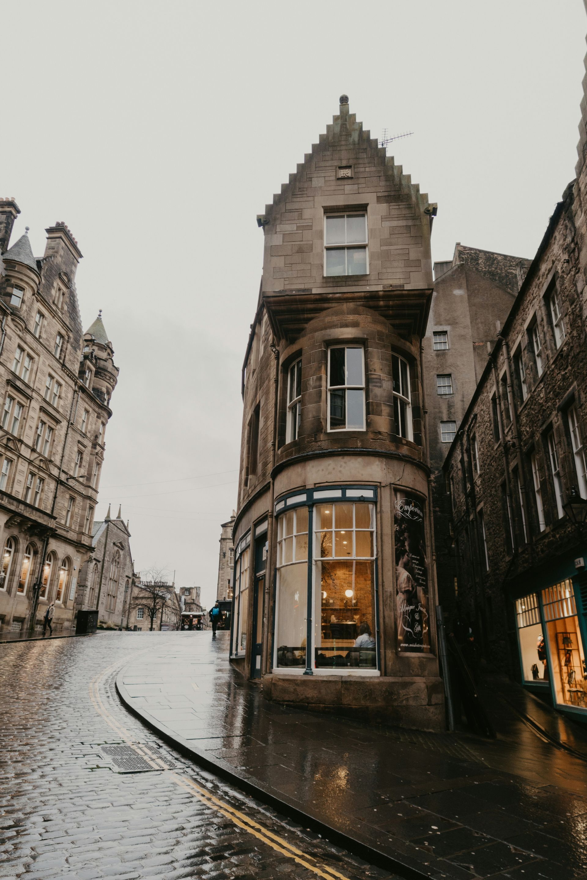 Stone buildings line a wet cobblestone street in Edinburgh. Brightly lit corner shop, overcast sky.