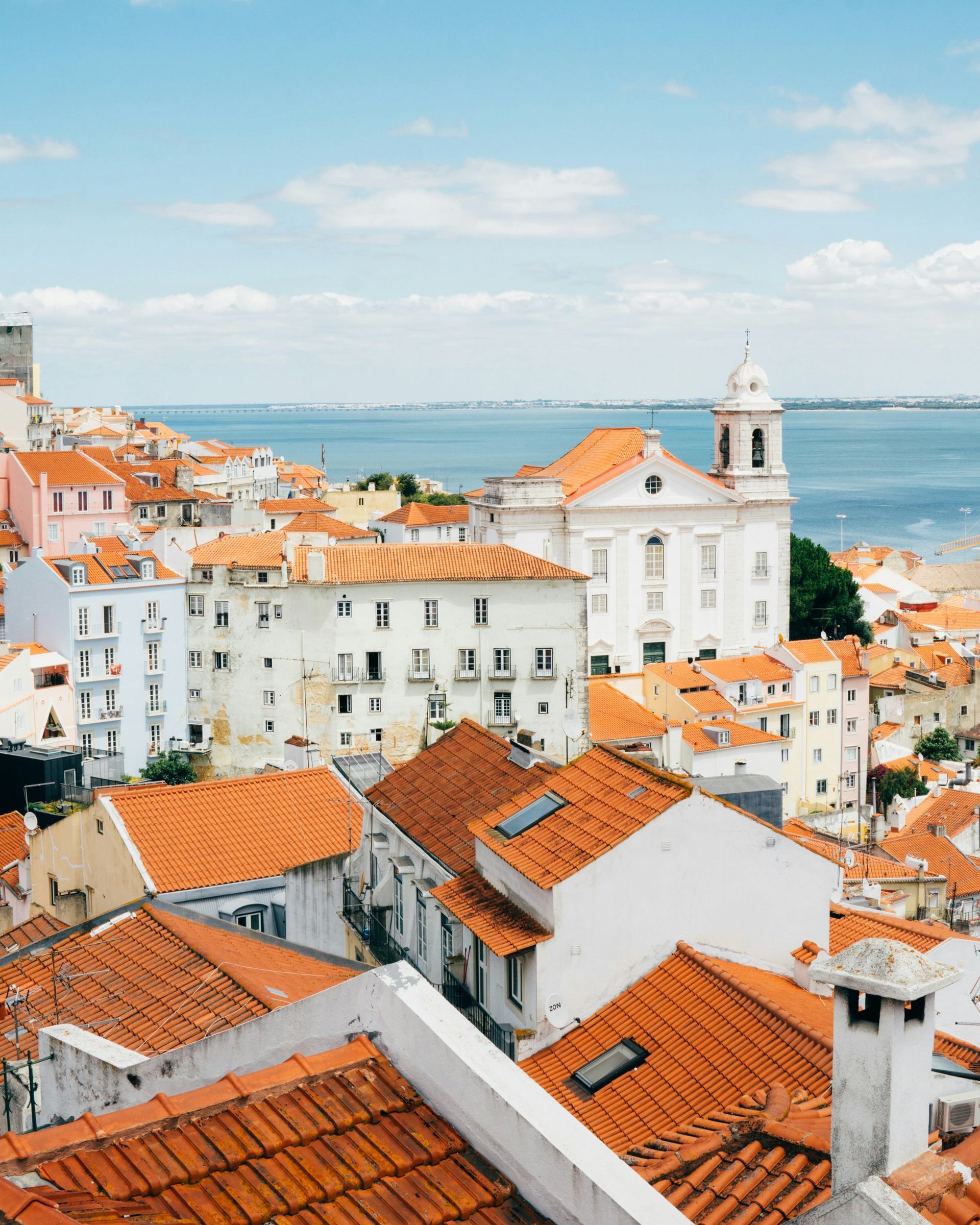 Cityscape of Lisbon, Portugal, with orange rooftops, white buildings, and a church against a blue sea.
