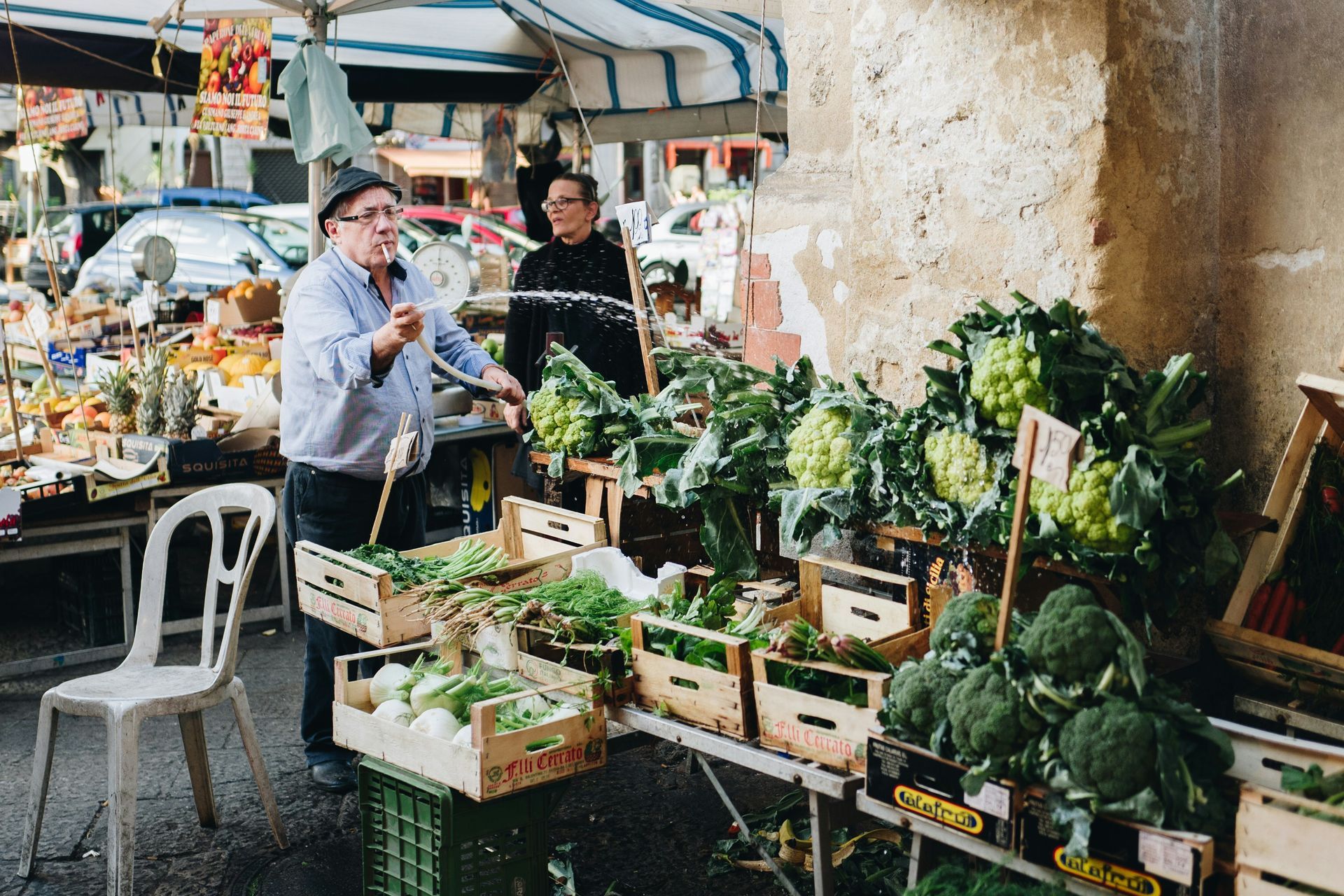 A man is standing in front of a vegetable stand at a market in Sicily.