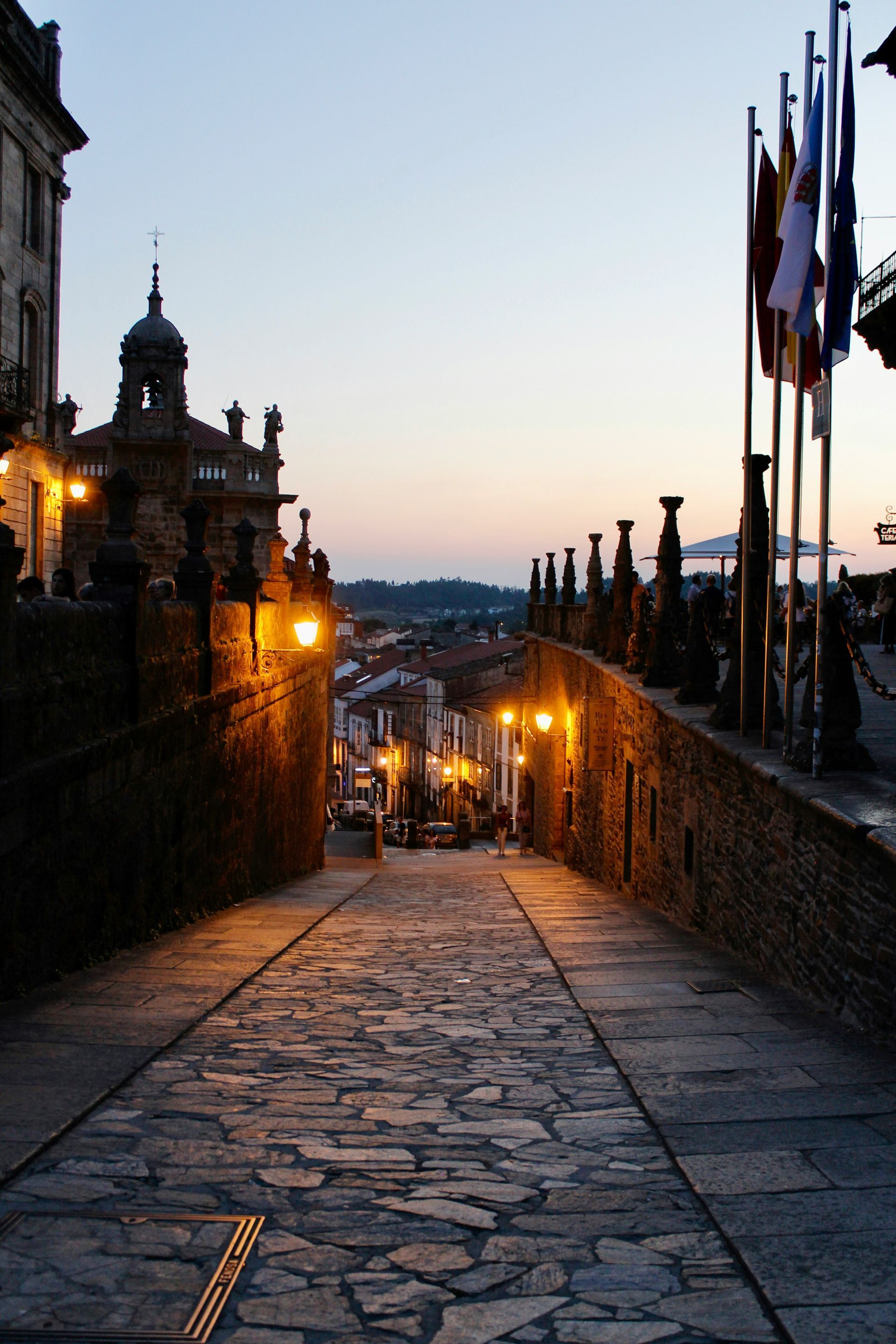 Cobblestone street lined with warm streetlights leading to a cityscape at dusk in Santiago de Compostela, Spain.