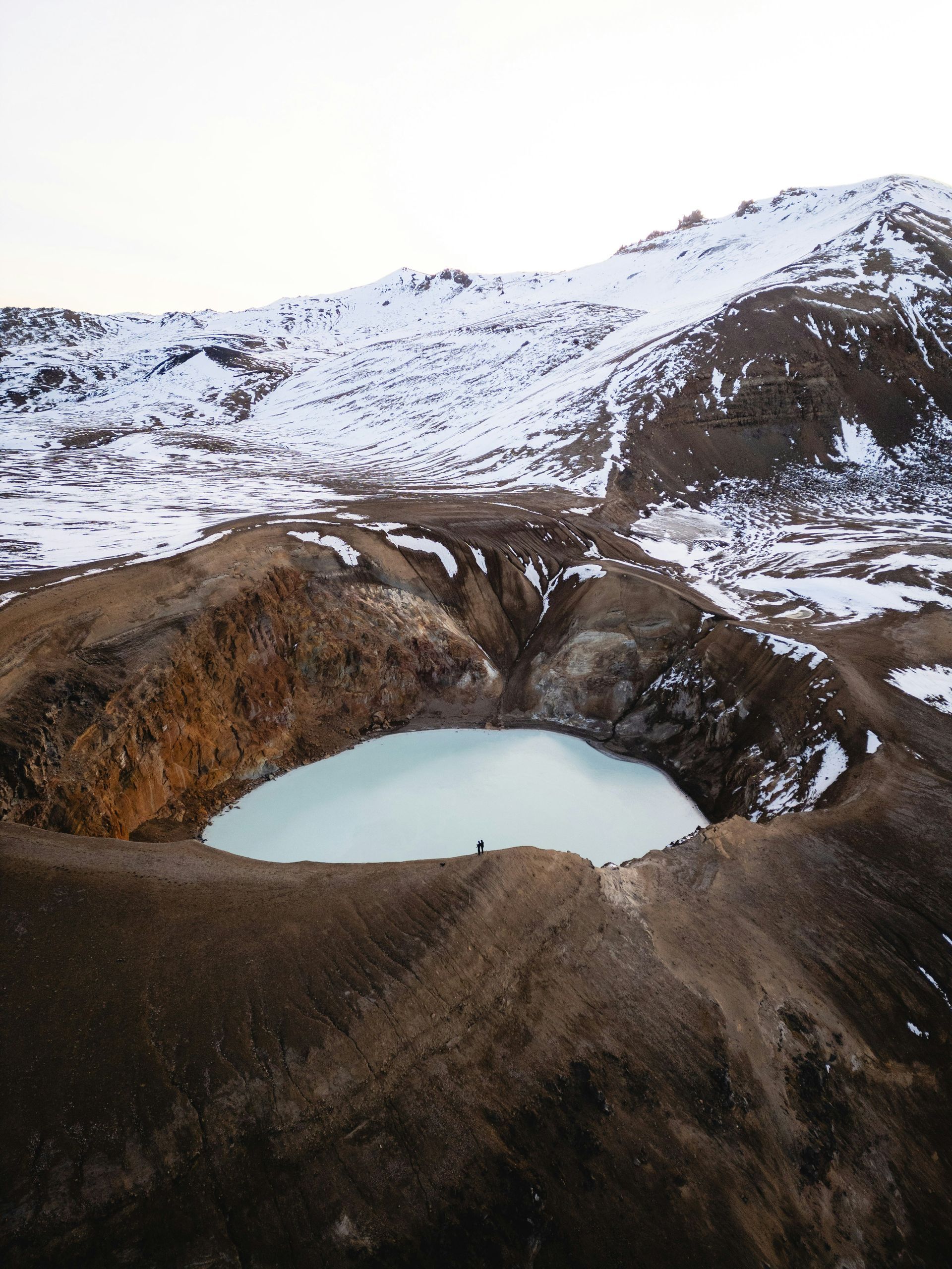 A person stands on the rim of a blue-water crater lake of Askja, surrounded by snowy brown mountains in Iceland.