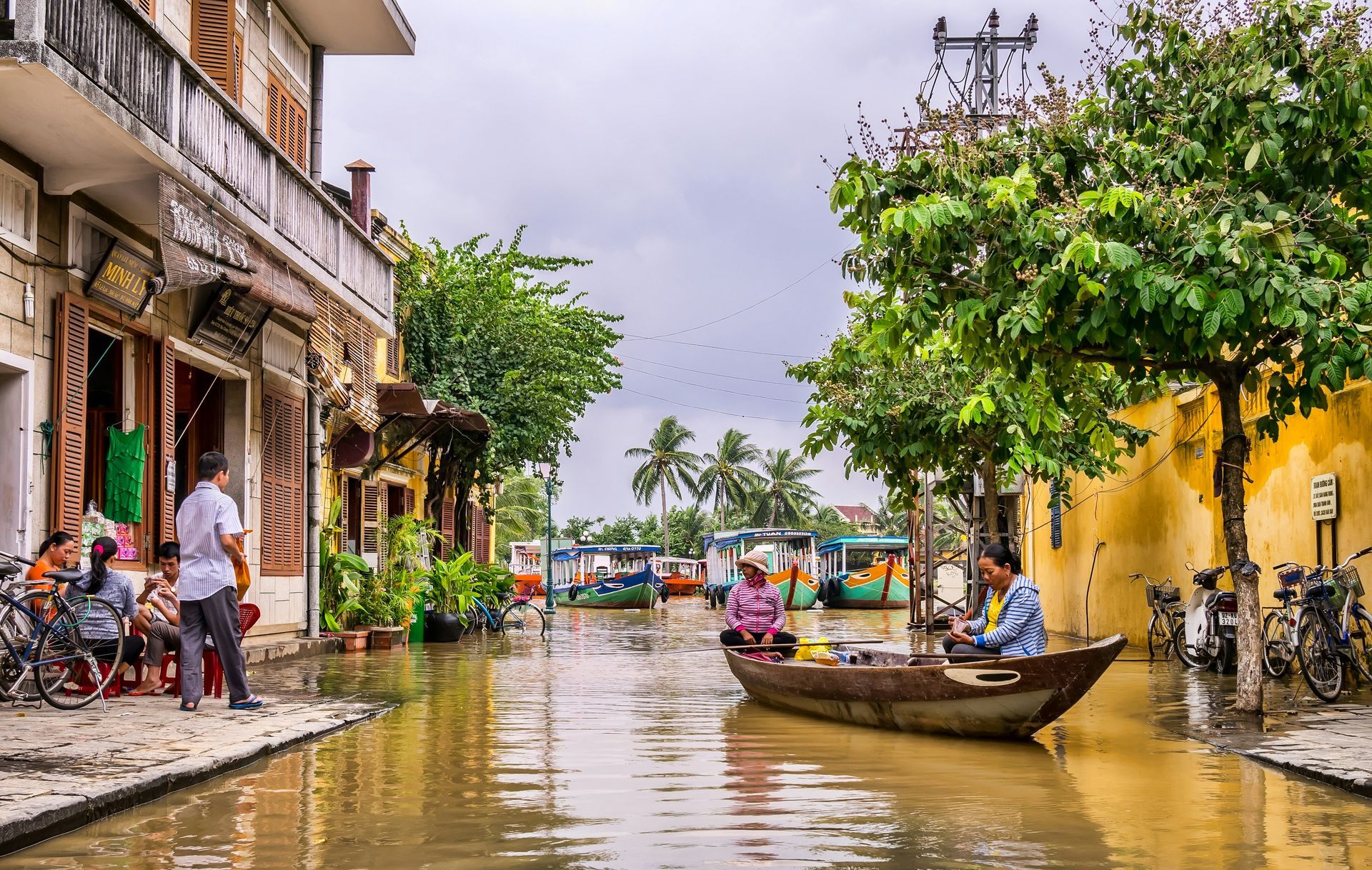 Flooded street in Hoi An, Vietnam. People in boats navigate between buildings. Brown water, green trees, and cloudy sky.
