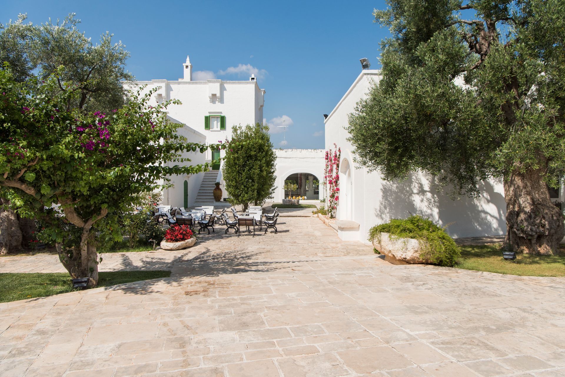 A white building with a stone driveway leading to it surrounded by trees and bushes at Il Melograno Hotel in Puglia, Italy.