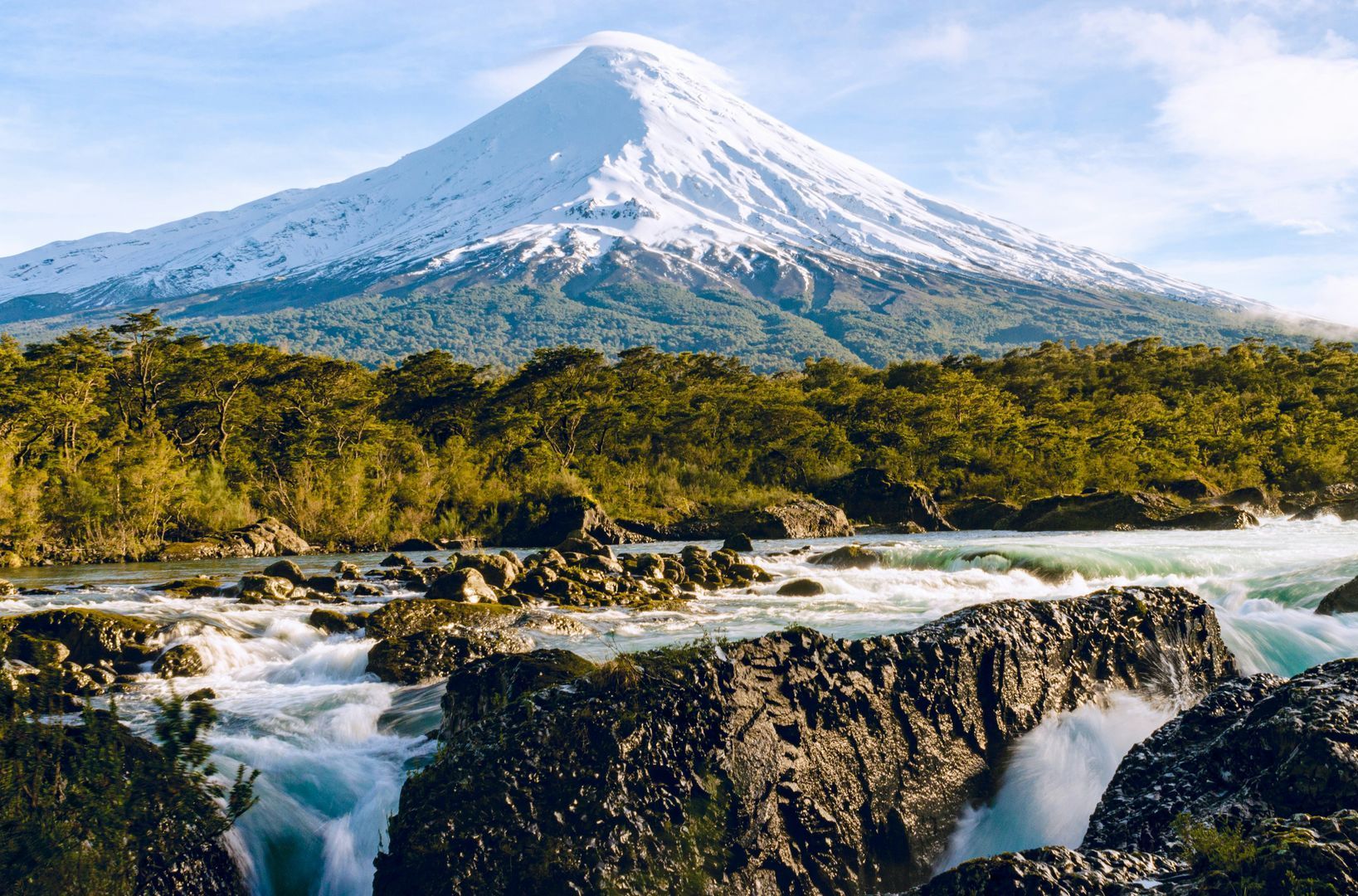 Snow-capped Osorno volcano overlooking a river with the Petrohué Waterfalls, surrounded by lush green trees in Chile.