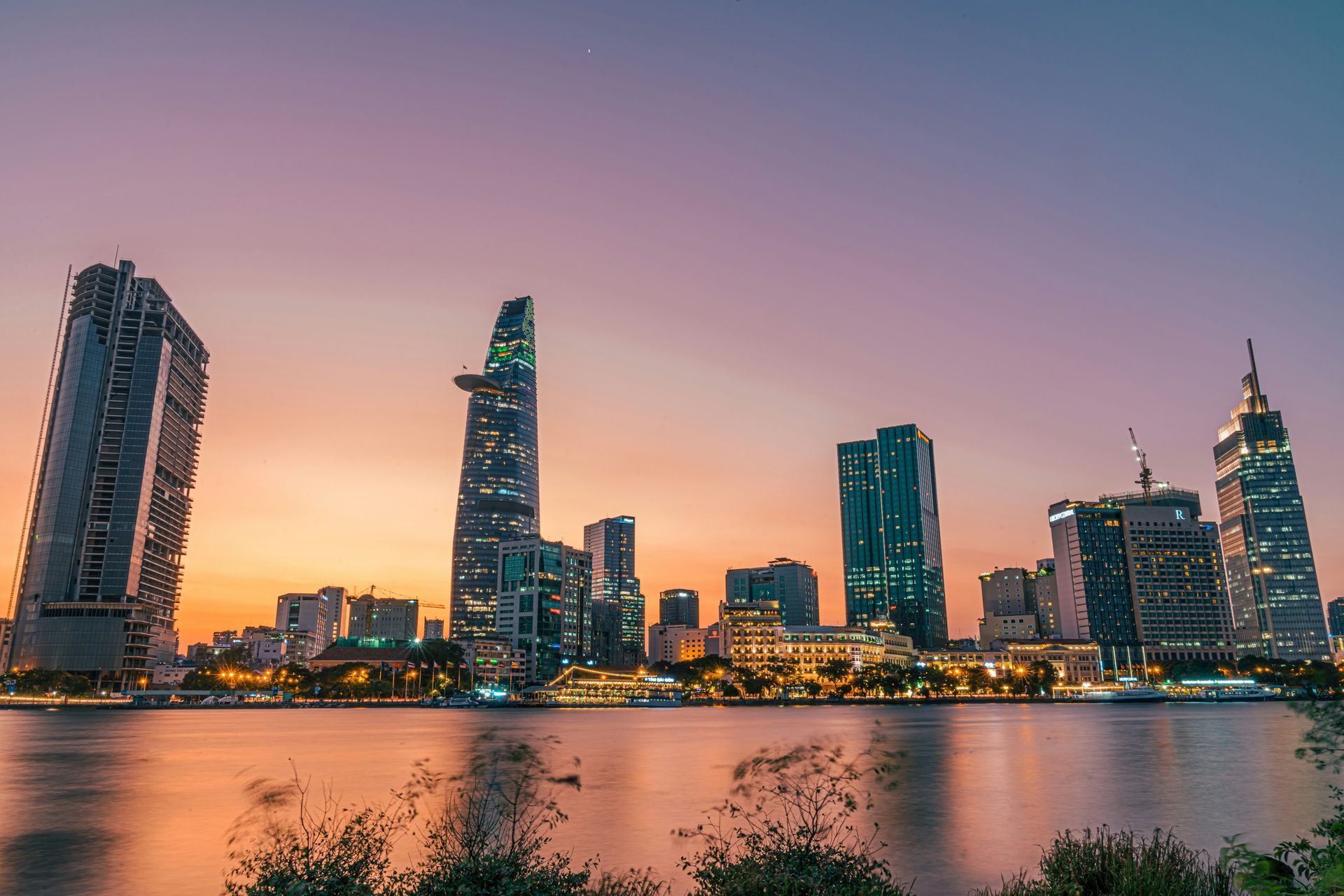 Skyline of Ho Chi Minh City at dusk, buildings silhouetted against a colorful orange and purple sky over water.