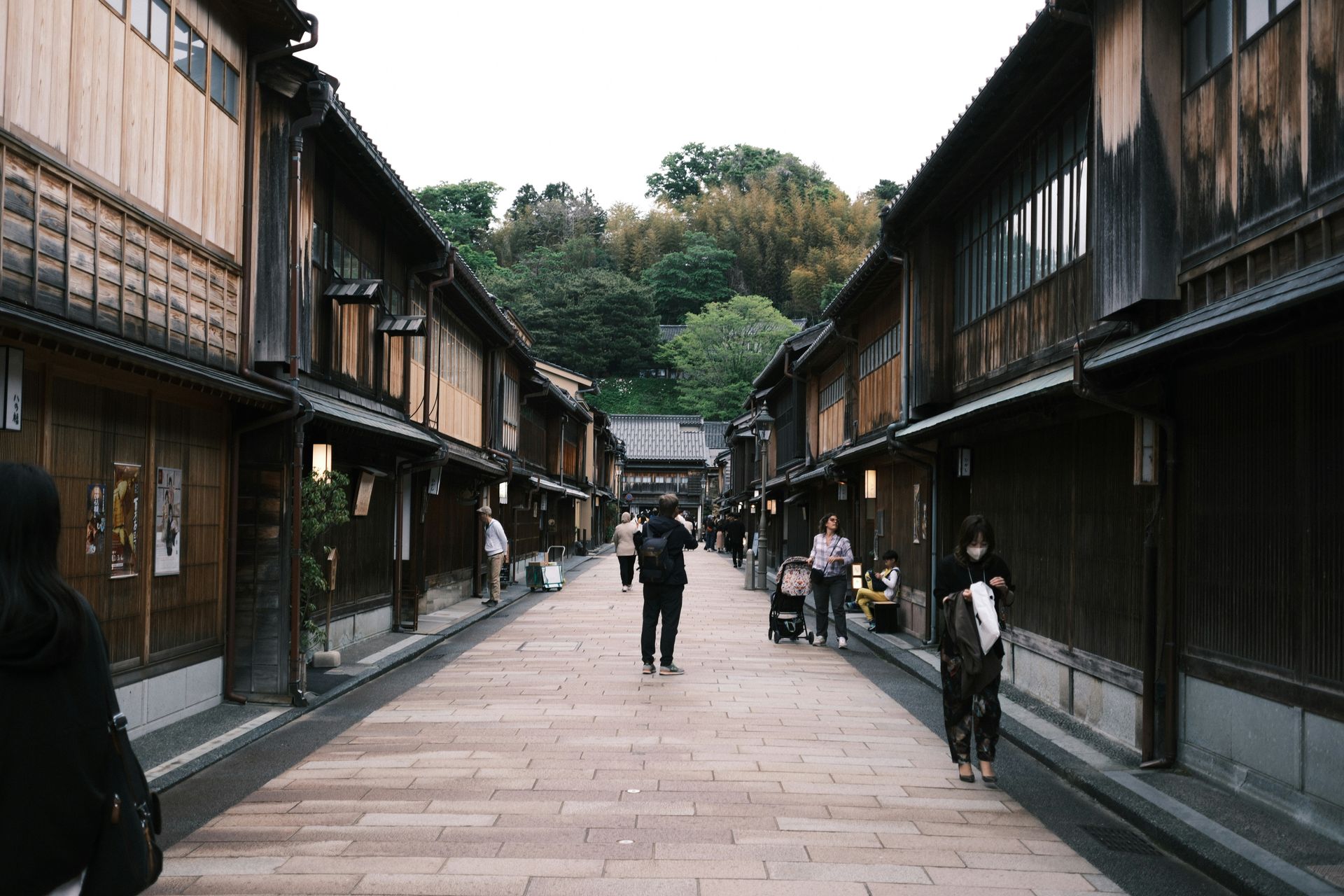 Street lined with wooden buildings in Kanazawa, Japan, pedestrians walking, cloudy sky.