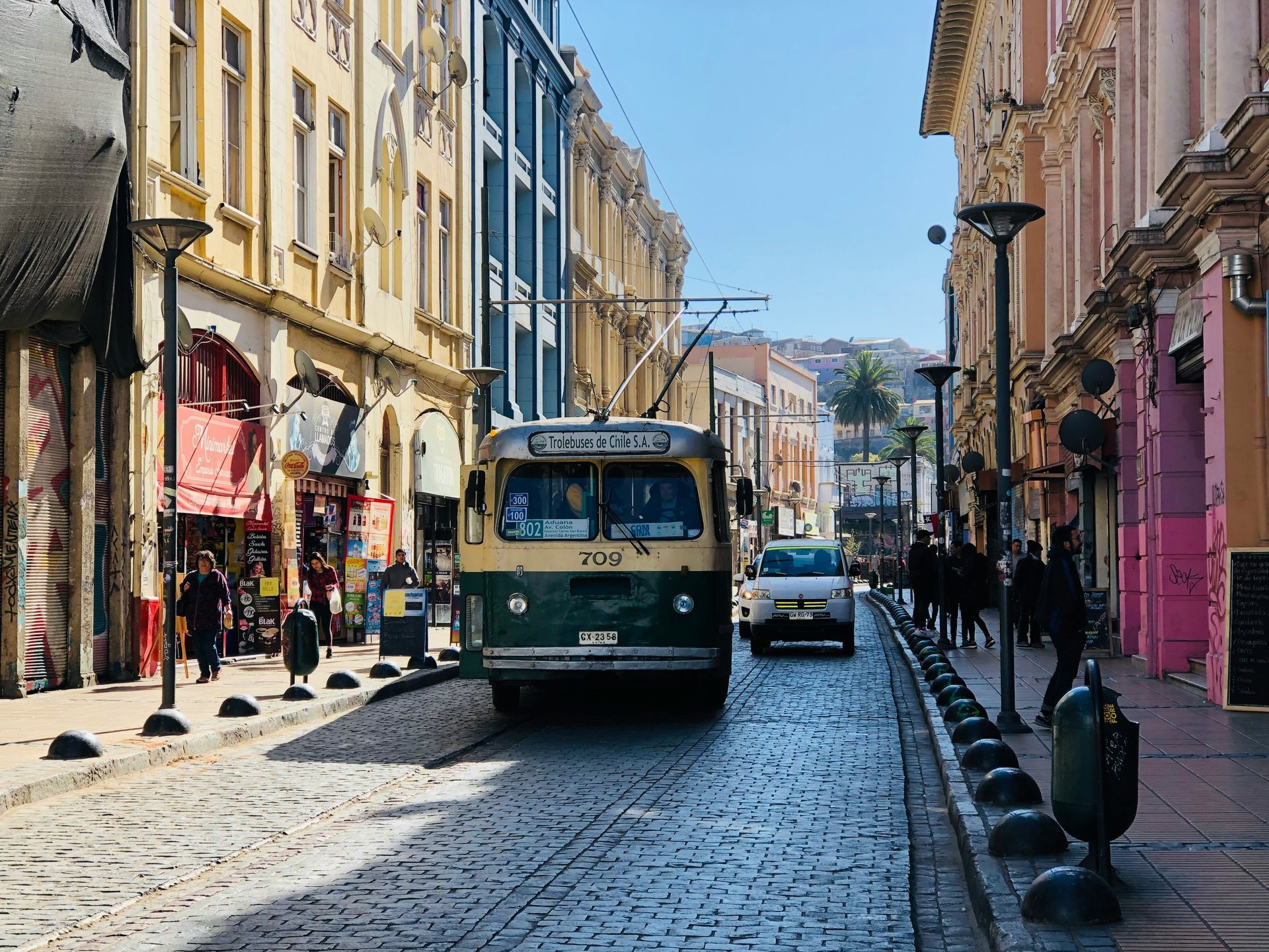 Vintage trolley bus on cobblestone street, between colorful buildings with people, in Valparaíso, Chile.