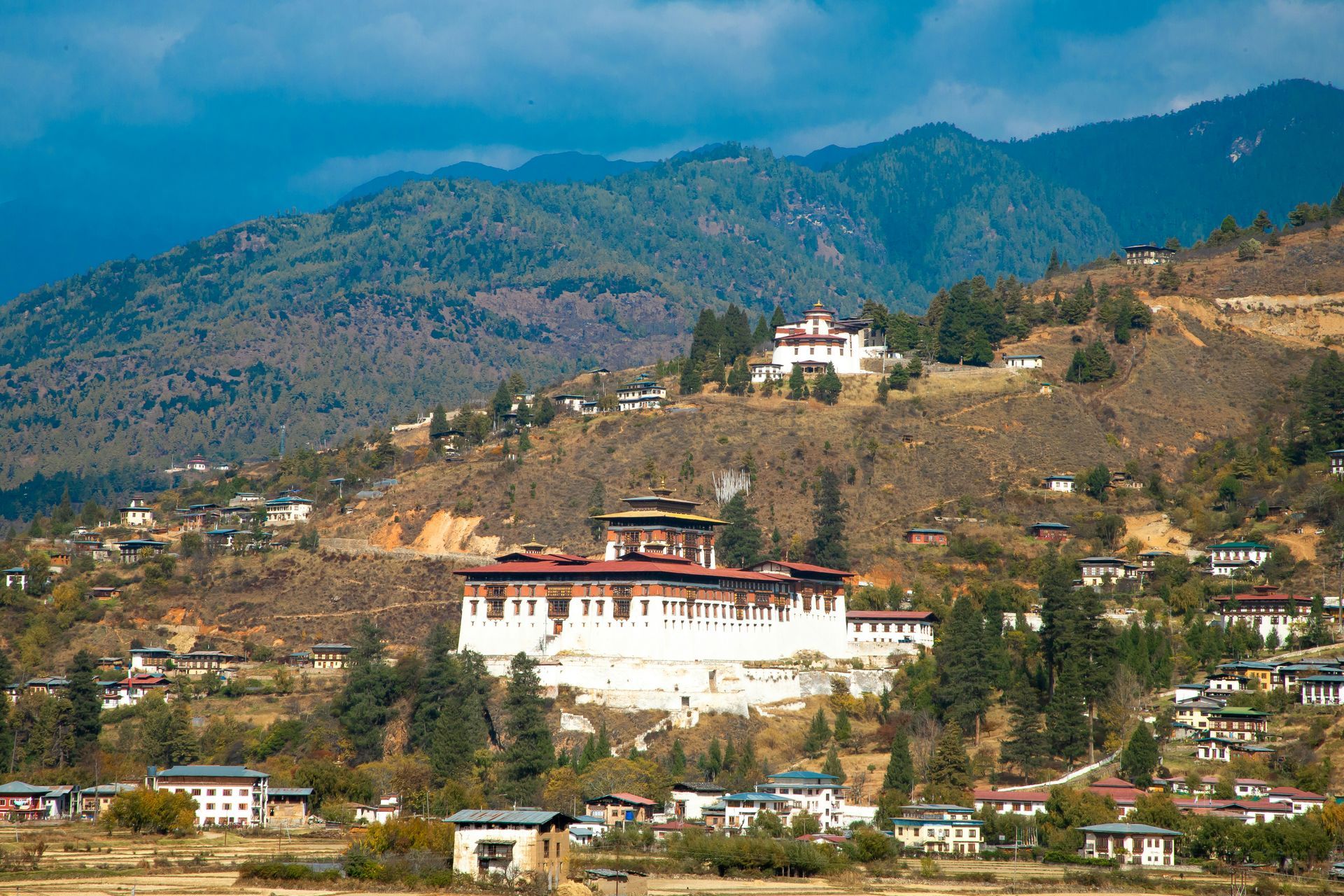 Rinpung Dzong with red and white walls sits on a hillside in Paro, Bhutan, surrounded by buildings and mountains.