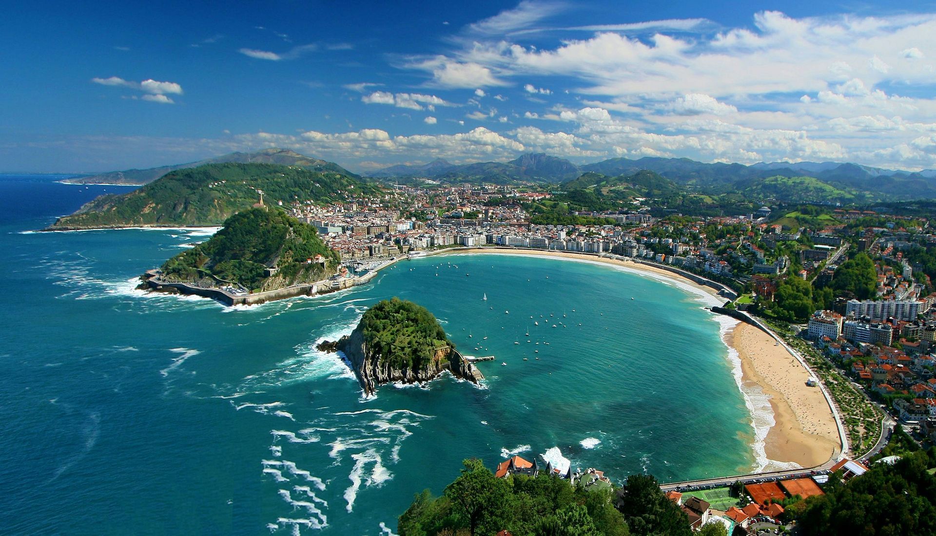 San Sebastián, Spain with a crescent-shaped beach, blue water, and lush green hills under a partly cloudy sky.