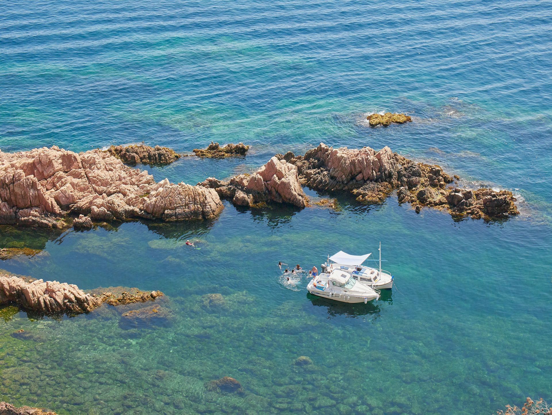 Clear turquoise water with a boat near reddish rocks. People swim around the boat on a sunny day on Costa Brava, Spain.