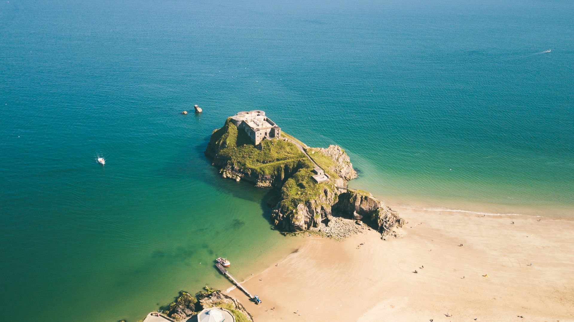 Island rock with ruins, sandy beach, and turquoise sea, seen from above in Tenby, Wales.