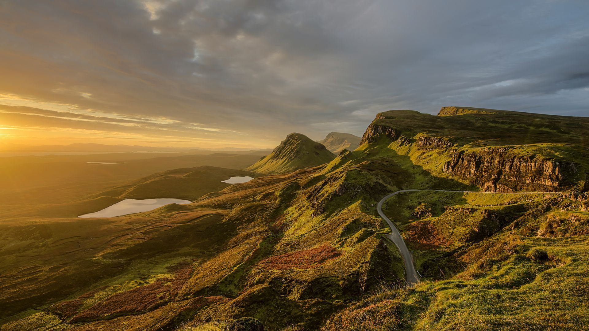 Golden sunrise over green, rocky mountains and a winding road; small lakes below on the Isle of Skye, Scotland.