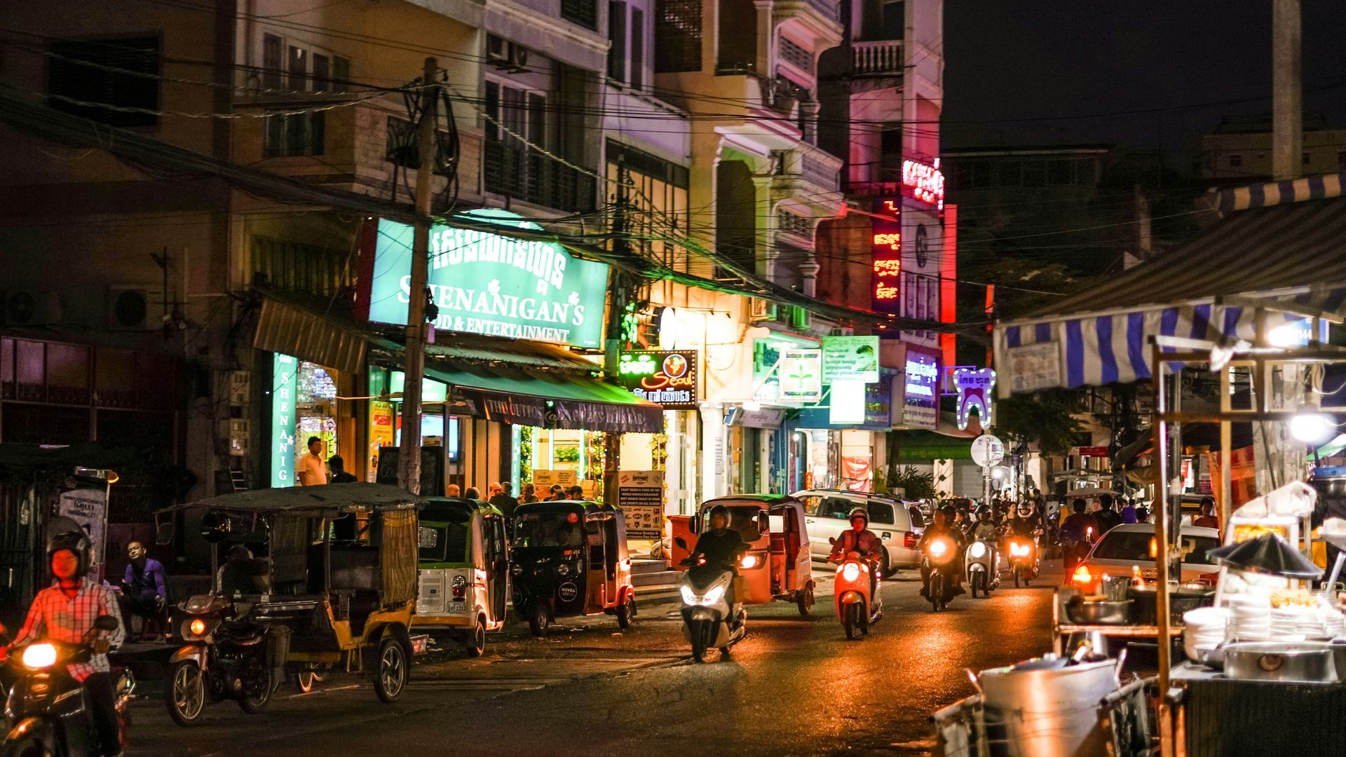 Nighttime street scene in Phnom Penh, Cambodia, with motorbikes, neon signs, and food stalls.