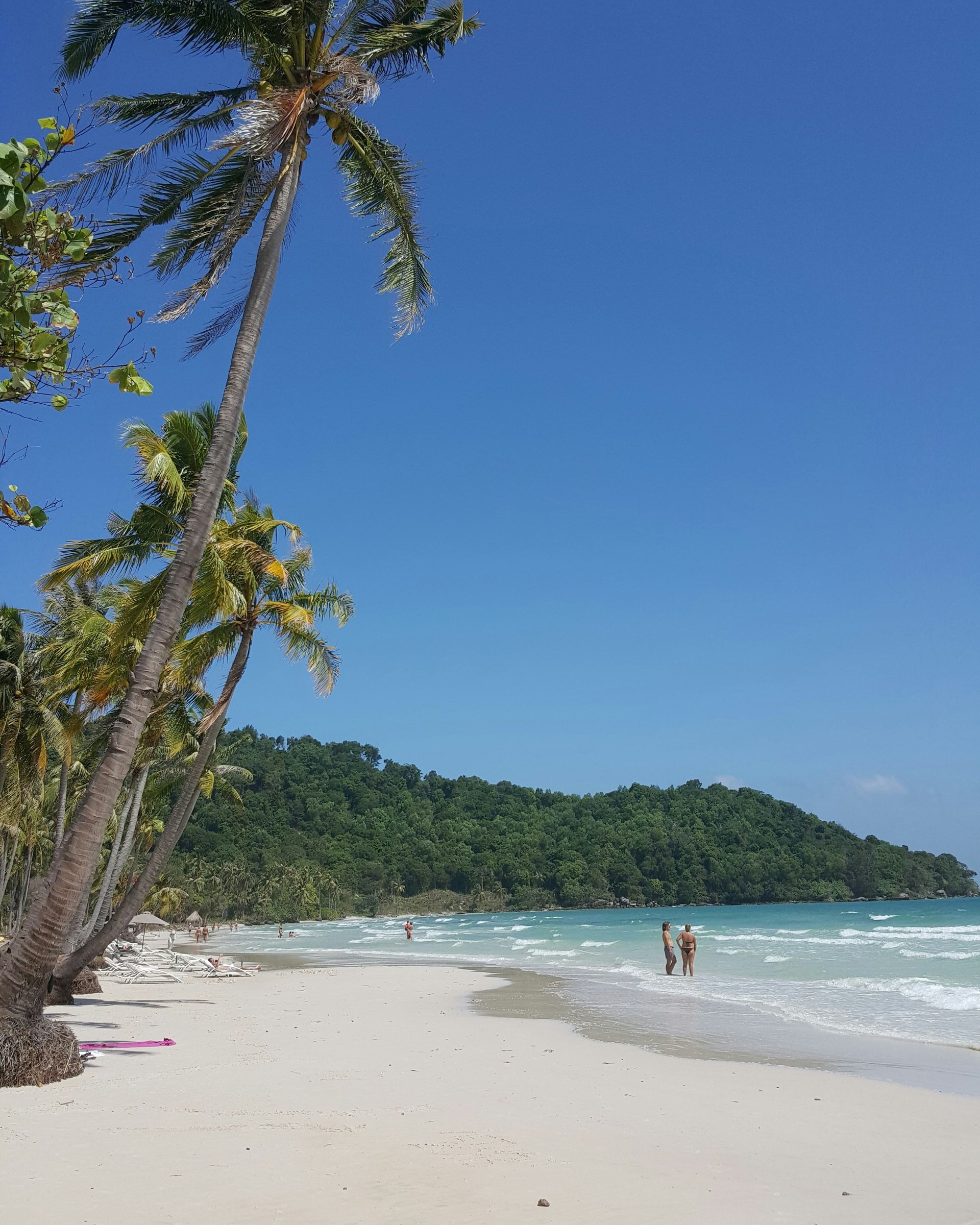 Palm trees frame a white sandy beach with turquoise water and a forested hill in Phú Quốc, Vietnam.