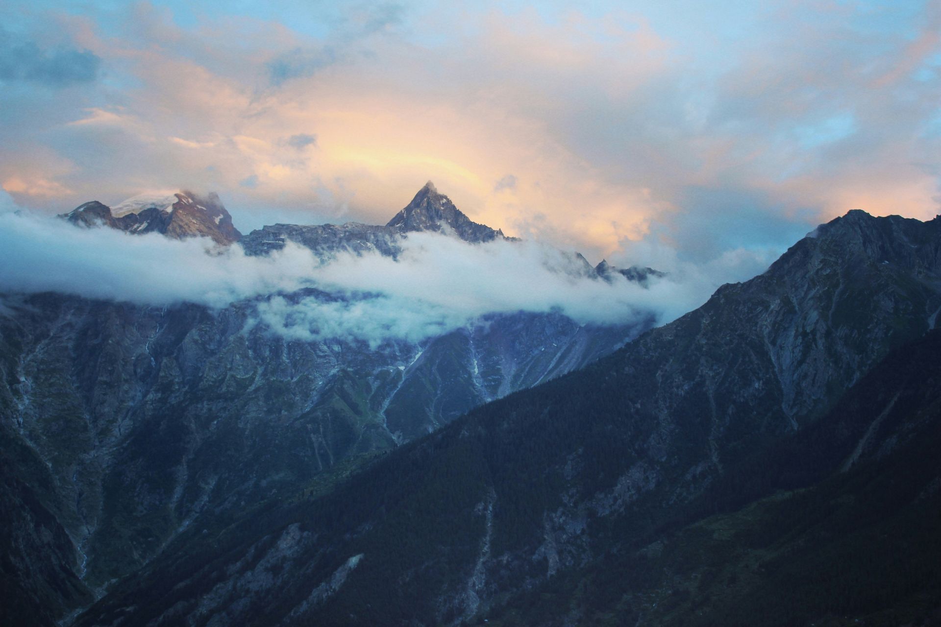 Mountains peak through clouds at sunset, sky with pink and gold hues in Ladakh, India.