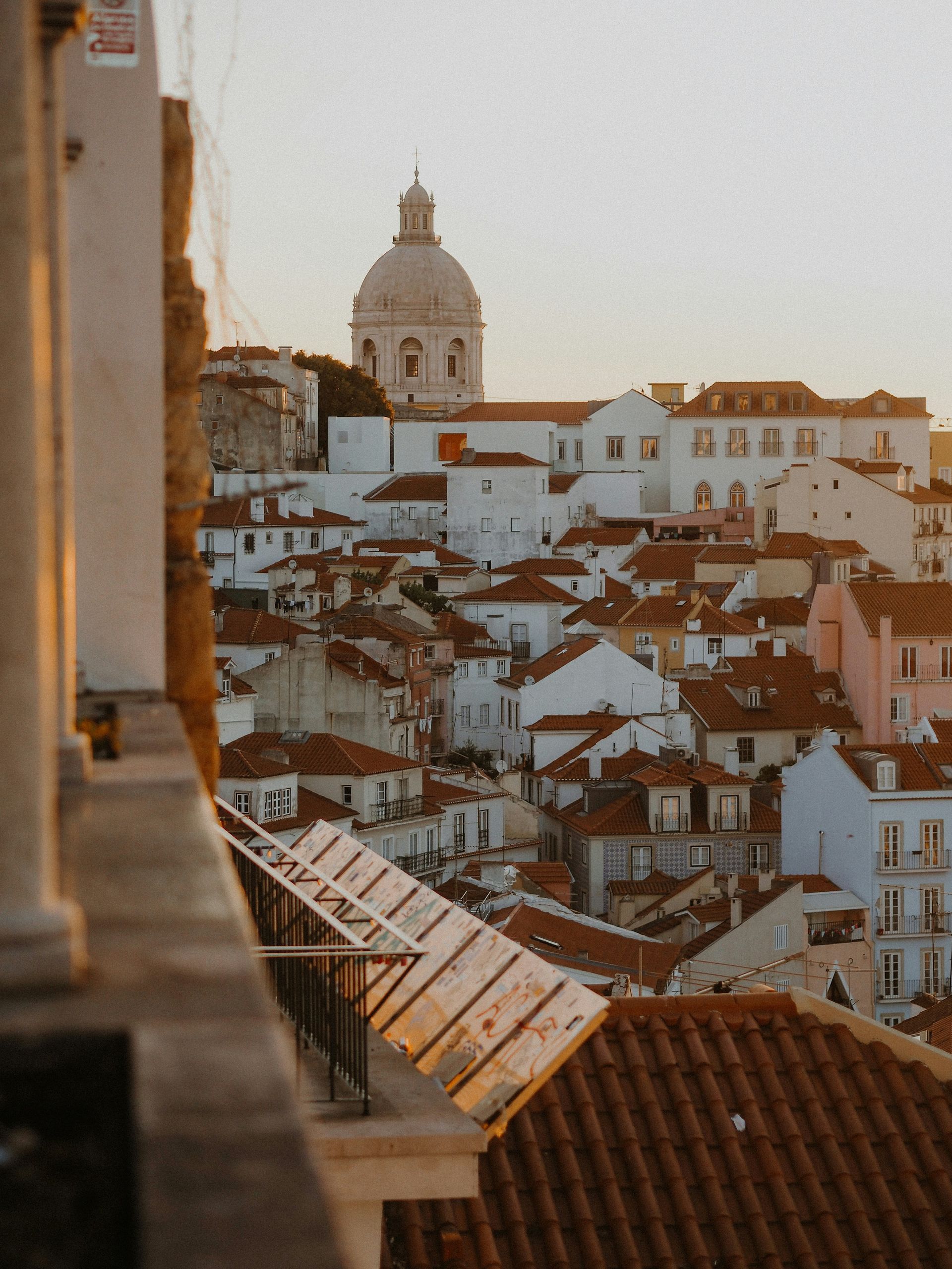 View of Lisbon, Portugal, with white buildings, red-tiled roofs, and a domed church under a warm sunset.