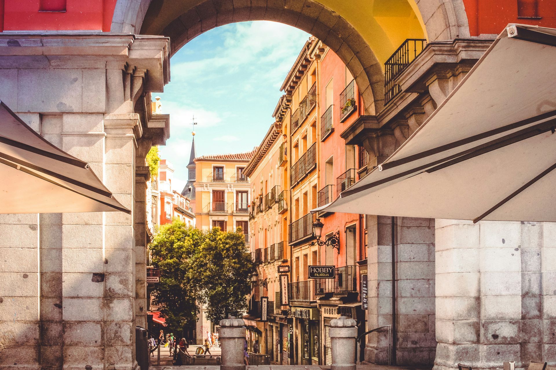 Stone archway frames a sunlit street with colorful buildings and outdoor cafe umbrellas in Madrid, Spain.