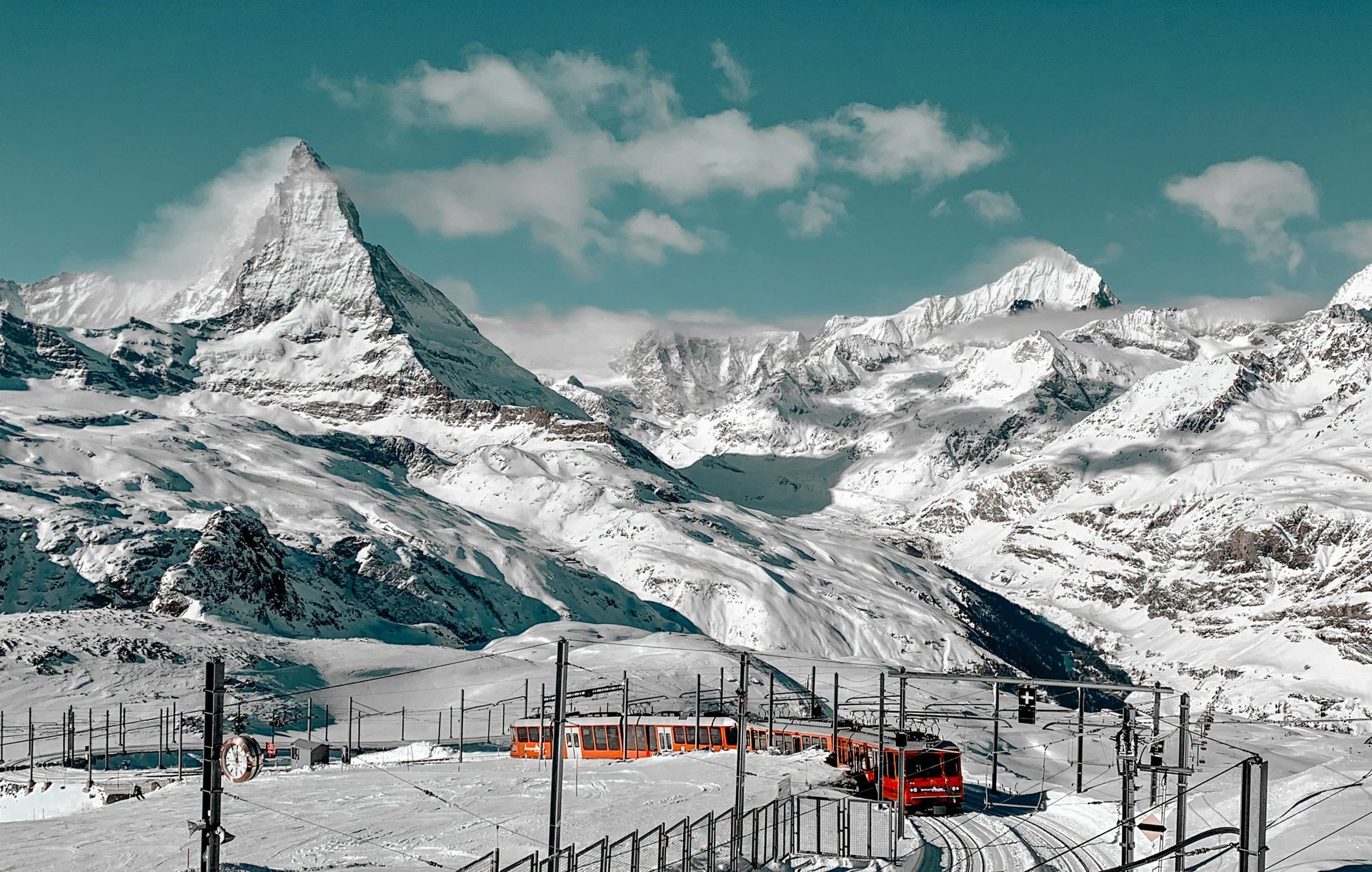 Snow-covered mountain range with a red train on tracks; bright blue sky, light clouds in Zermatt, Switzerland.