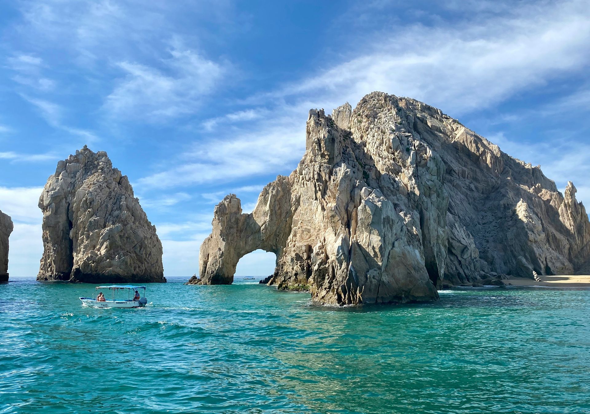 Rocky arch and formations rise from turquoise water under a blue sky. A small boat floats nearby in Cabo San Lucas, Mexico.