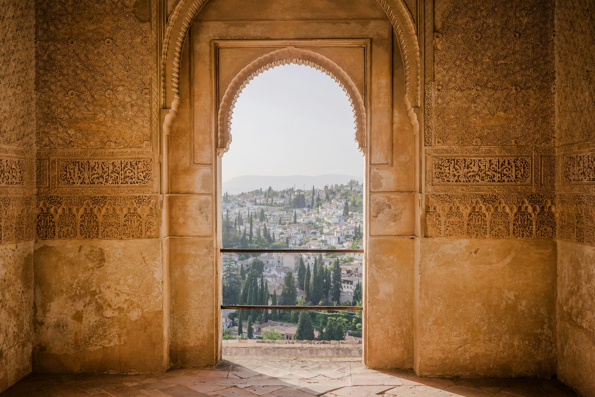 Ornate archway frames a view of trees and mountains. Warm-toned walls with detailed carvings in Granada, Spain.