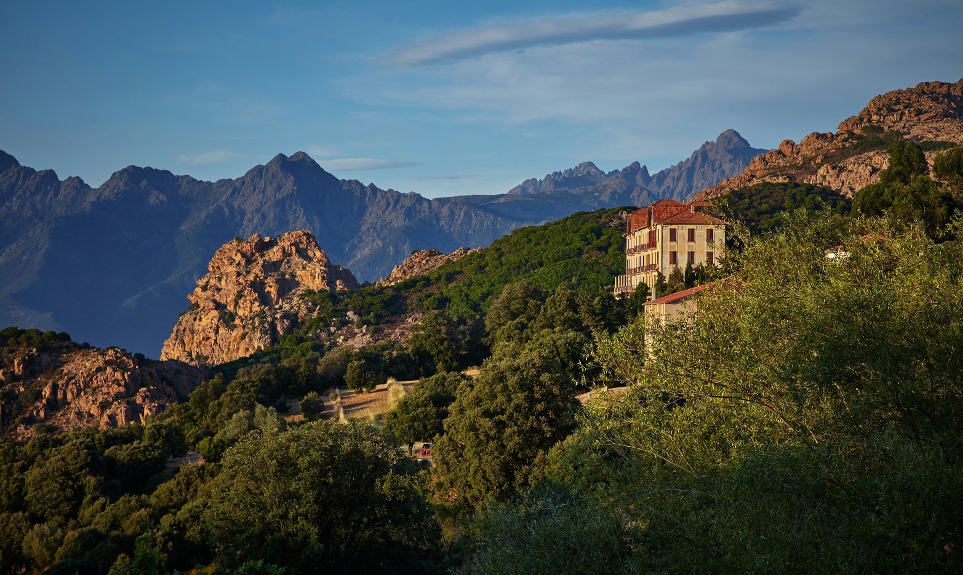 A house is sitting on top of a hill with mountains in the background in Corsica, France.