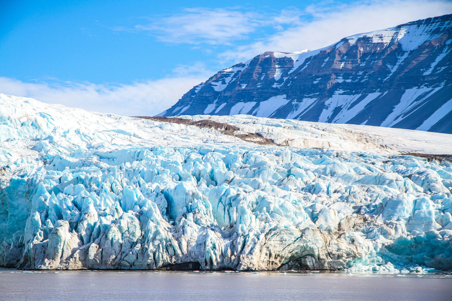 Glacier meets ocean, beneath a mountain range, bright blue and white in Svalbard, Norway.