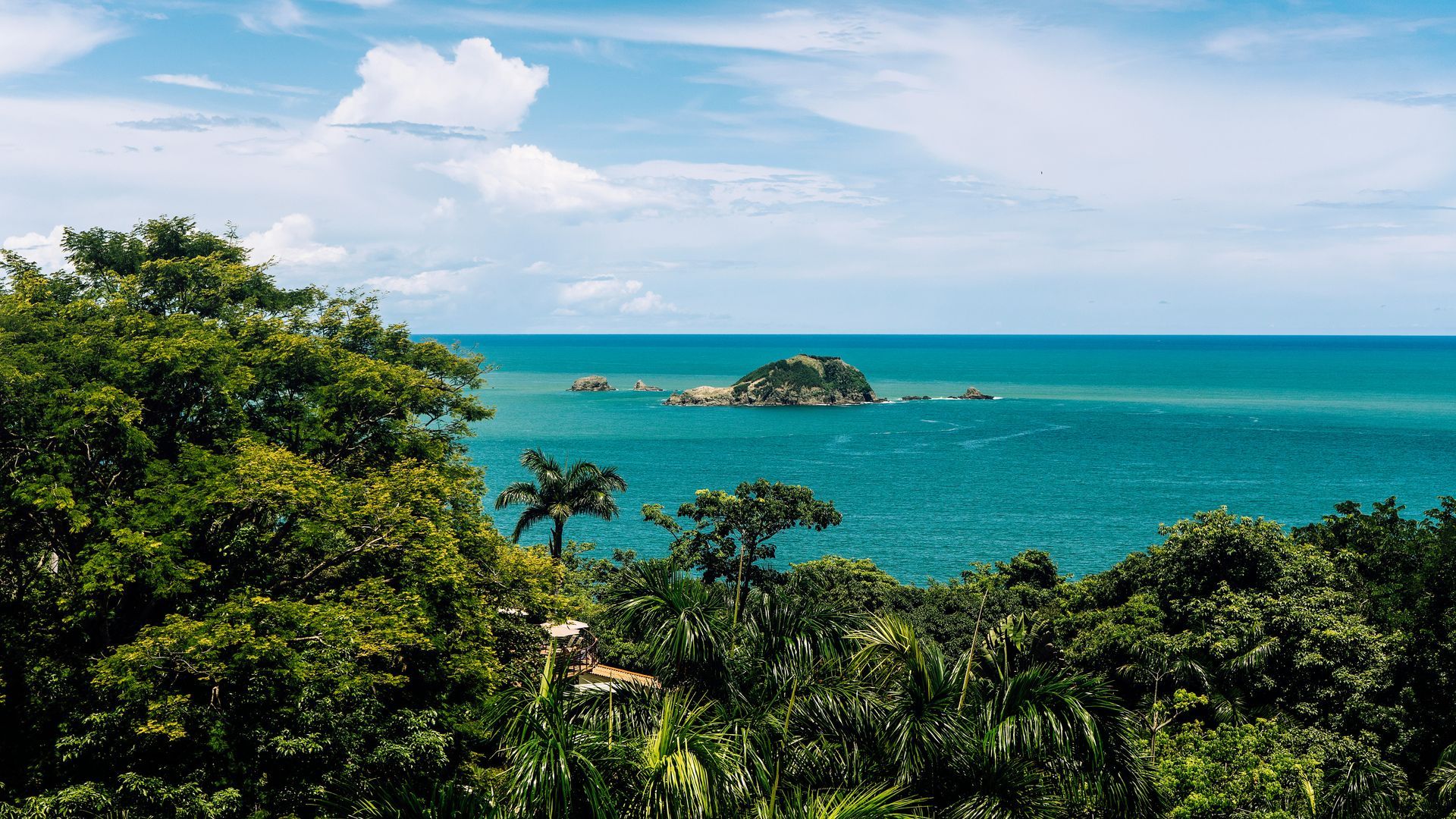 Vibrant blue ocean view with small island, framed by lush green trees under a partly cloudy sky in Manuel Antonio, Costa Rica.
