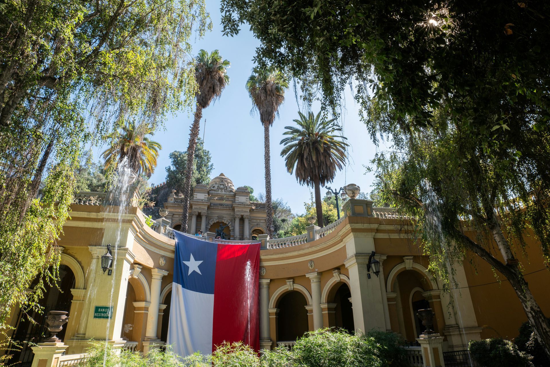 Chilean flag draped on a yellow building with archways, palm trees, and a fountain in Santiago, Chile.