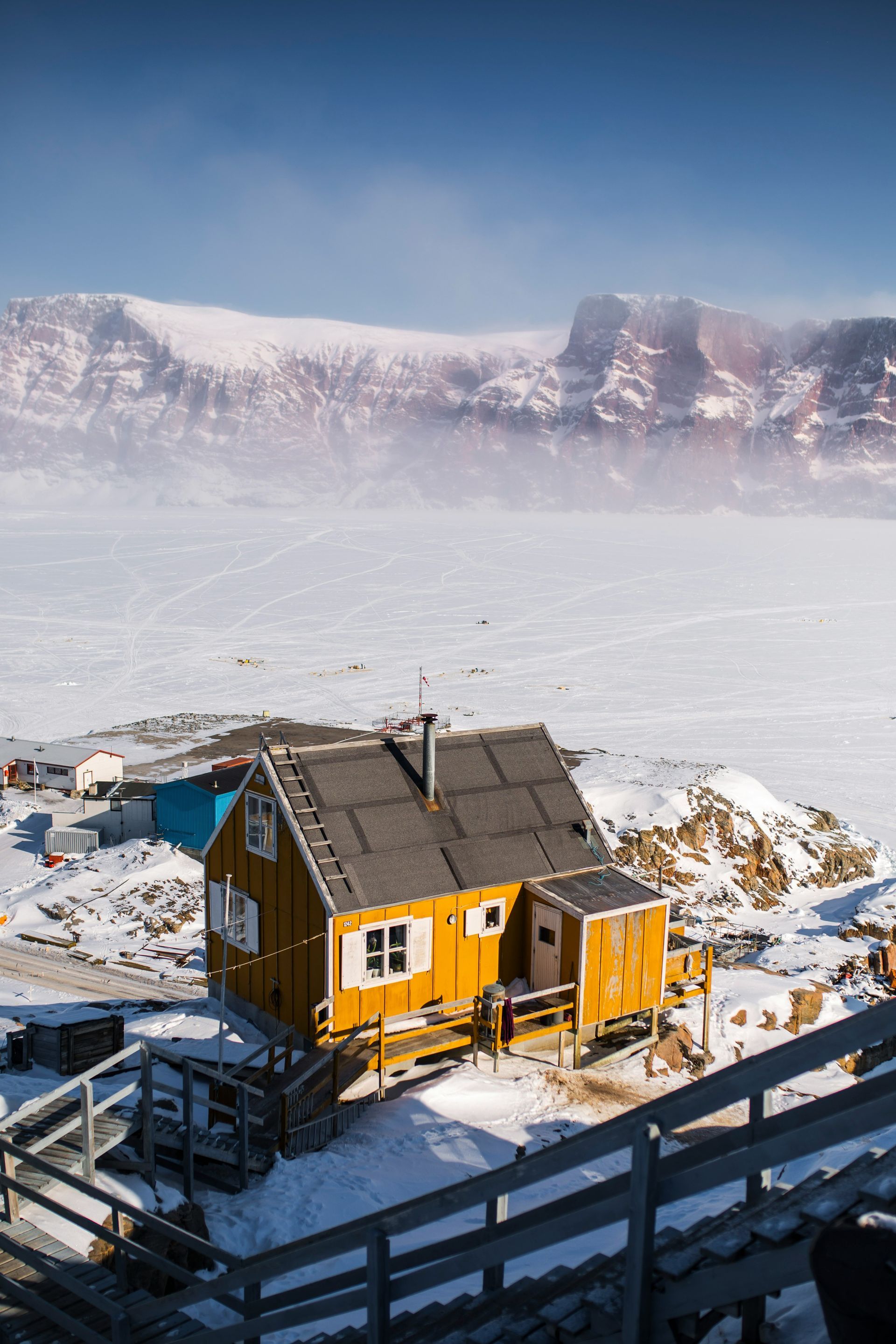 Yellow wooden house on a snowy cliff, overlooking an icy landscape, mountains in the background in Uummannaq, Greenland.