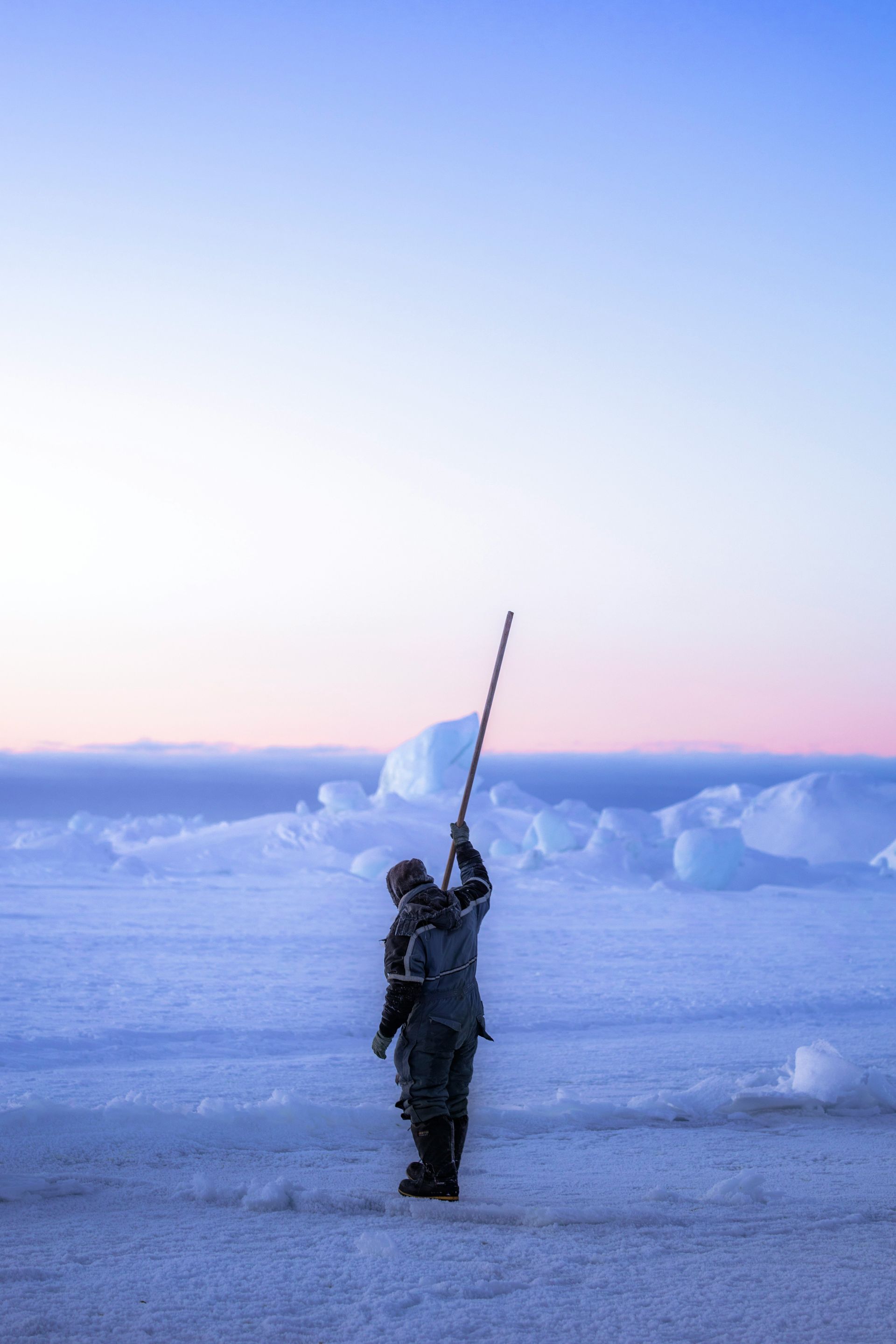 Person on a snowy plain holds a pole toward a blue and pink sky, near ice formations at the Ilulissat Icefjord, Greenland.