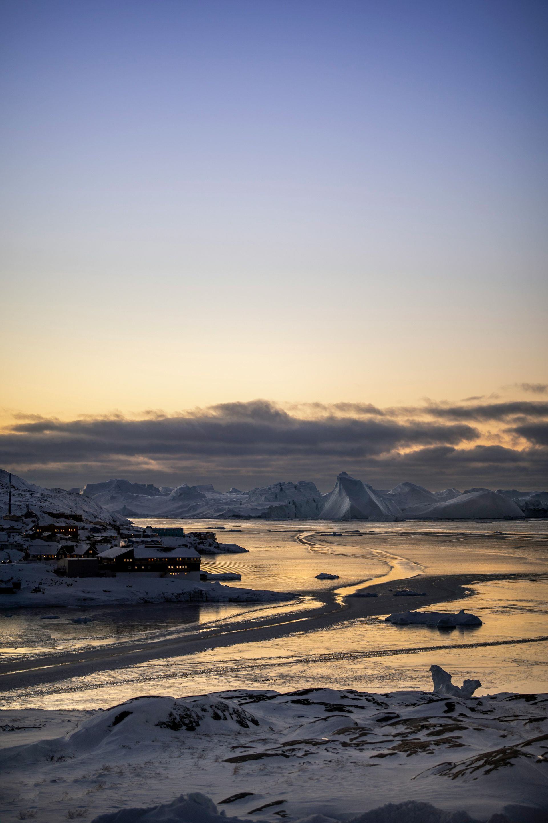 Snowy coastal town at dusk, with mountains and ice. Blue and orange hues in sky at the Ilulissat Icefjord, Greenland.