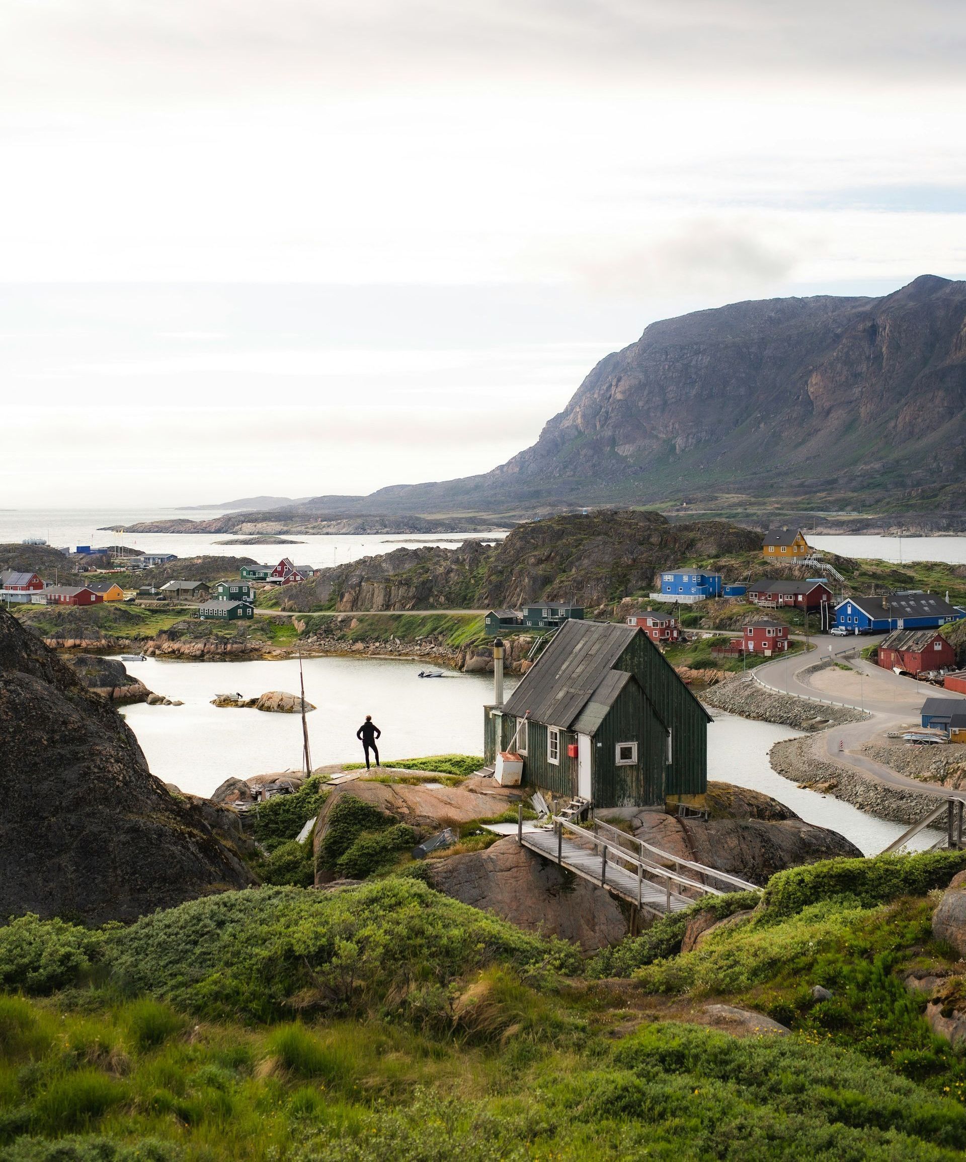 A person stands near a green church in the town of Sisimiut, Greenland, with colorful houses and mountains in the background.