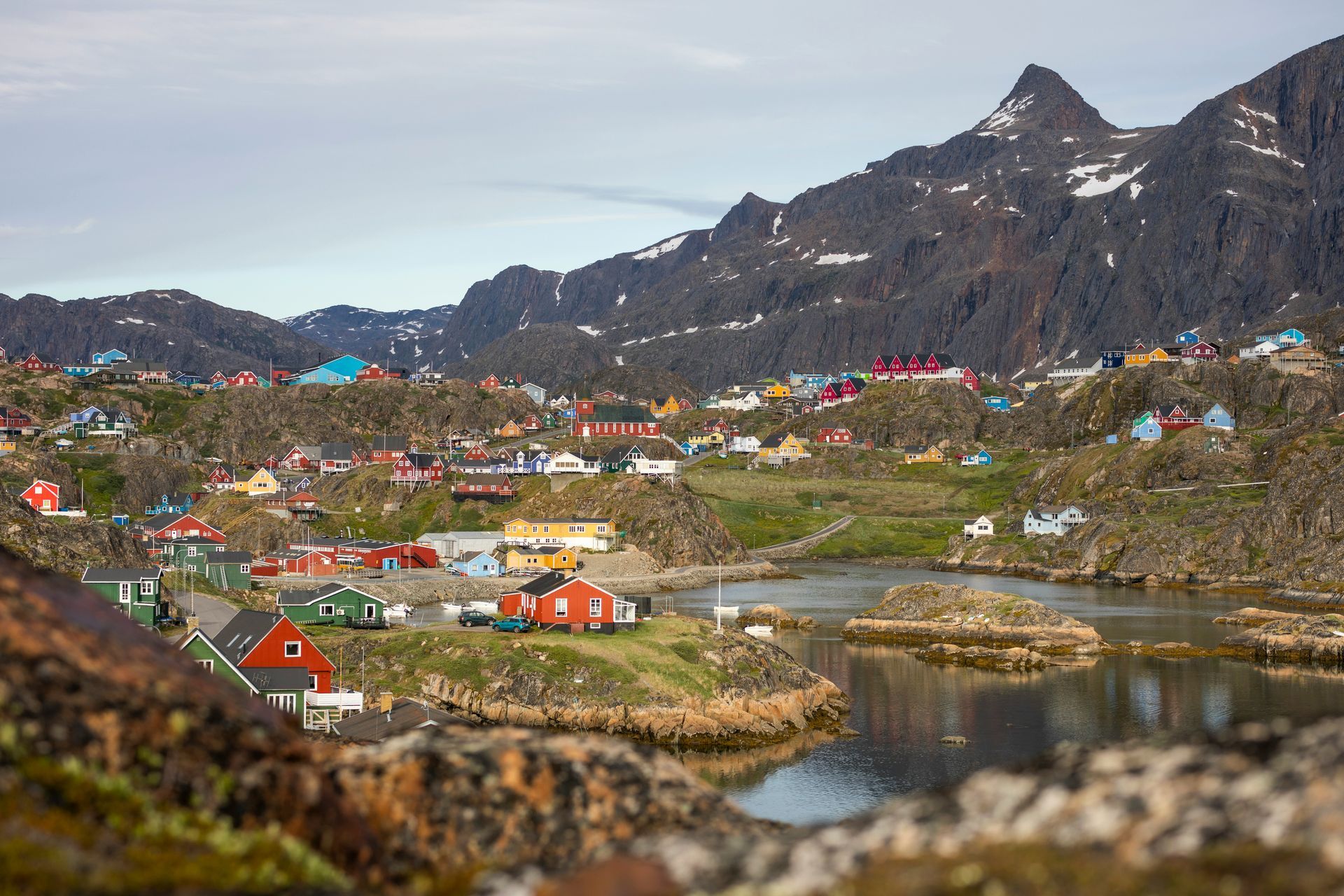 Colorful houses dot a rocky landscape in Sisimiut, Greenland, with mountains in the background and calm water in the foreground.