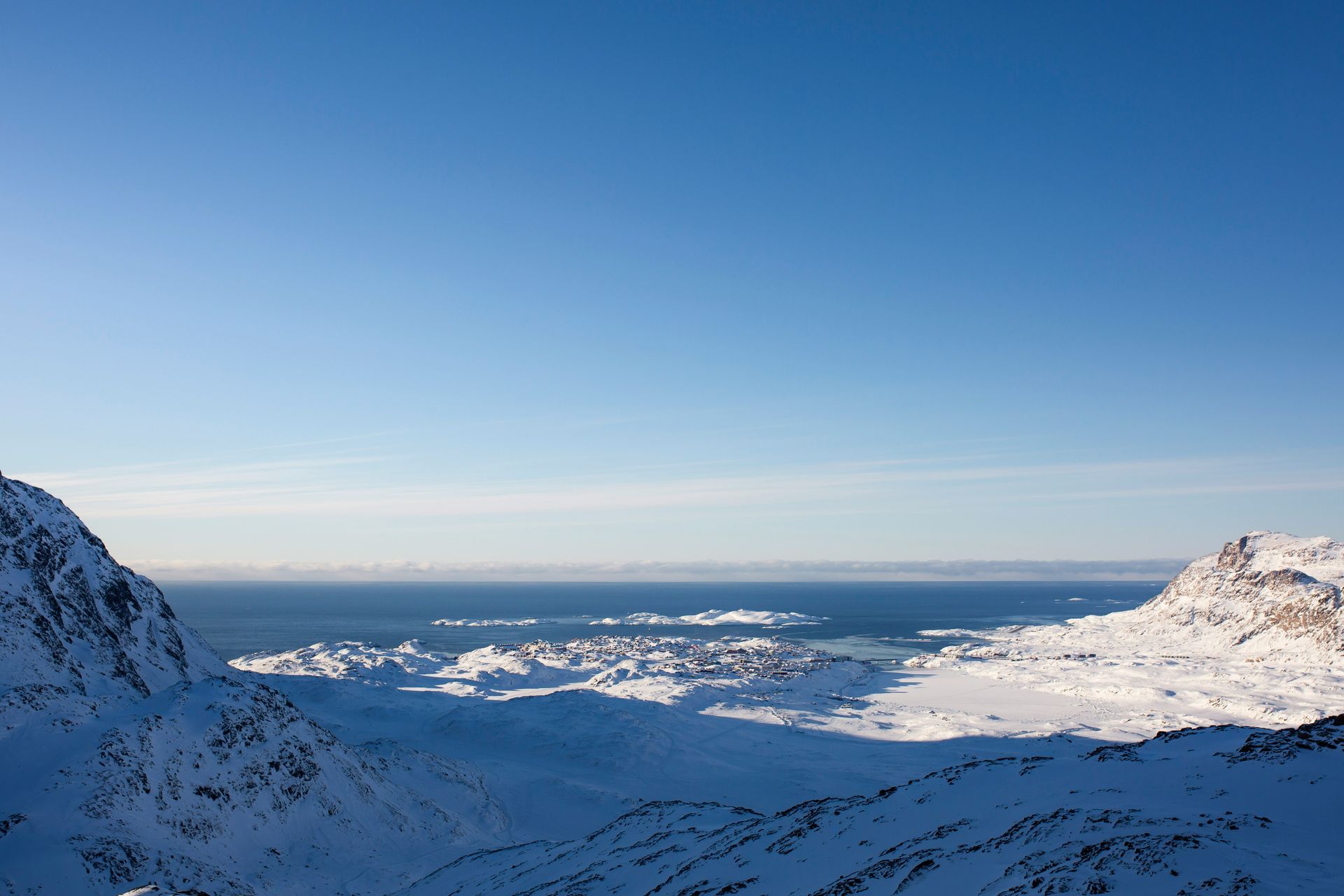 Snowy mountains overlook a coastal town, with a blue sky and ocean in the background in Sisimiut, Greenland.