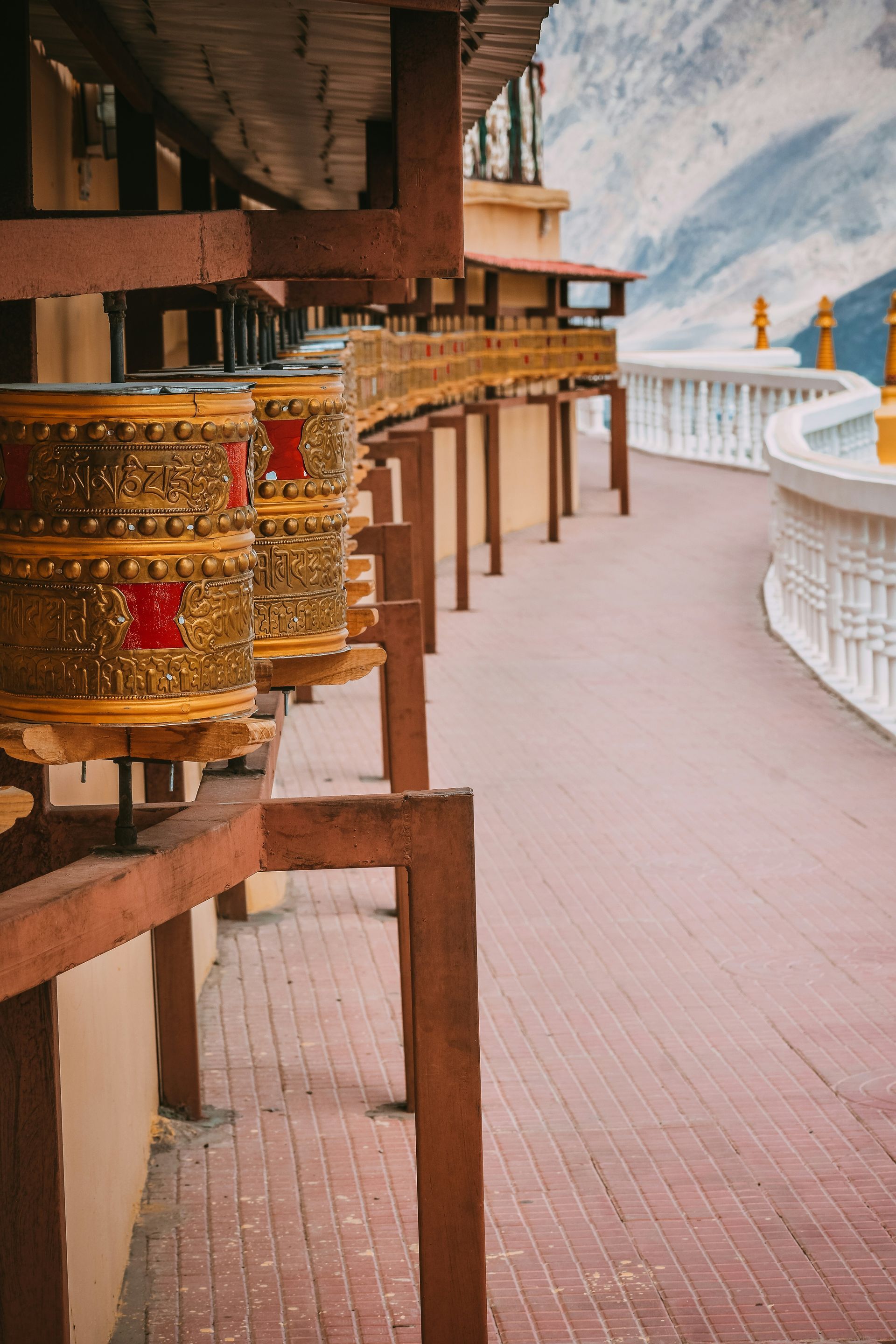 Golden prayer wheels along a brick walkway at a temple, with mountains in Ladakh, India.
